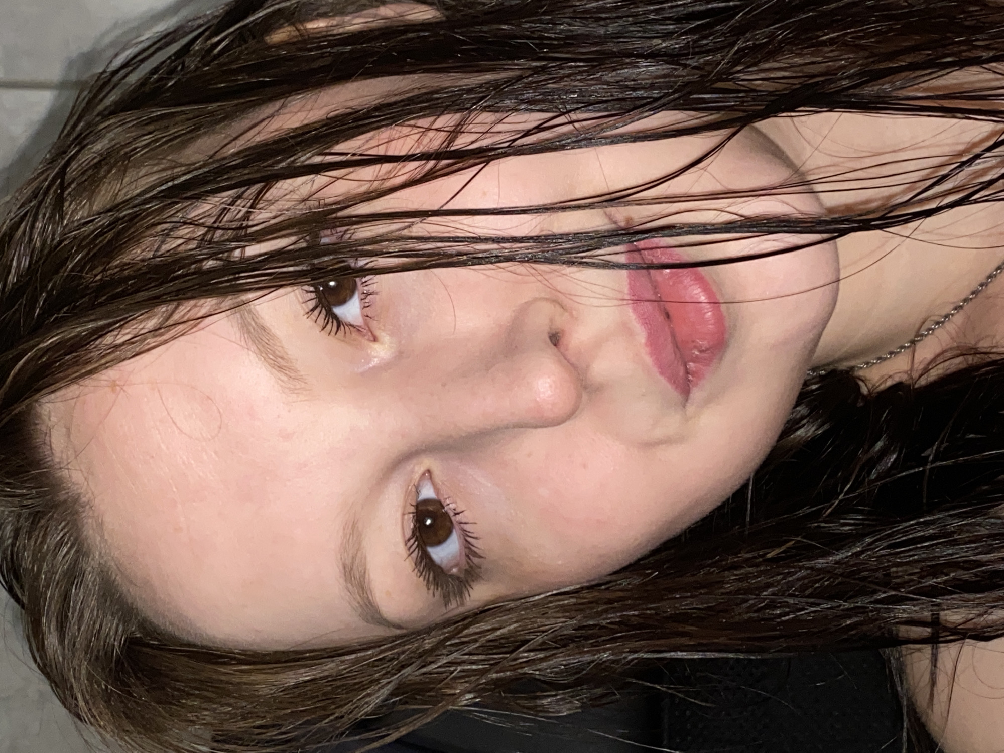 A close-up, angled portrait shows a fair-skinned person with wet, dark brown hair partially obscuring their face. The individual's eyes are brown, and their lips are a mauve color. A thin silver necklace is visible on their neck. The background is out of focus and indistinct. The location is not discernible from the visual information provided in the image.