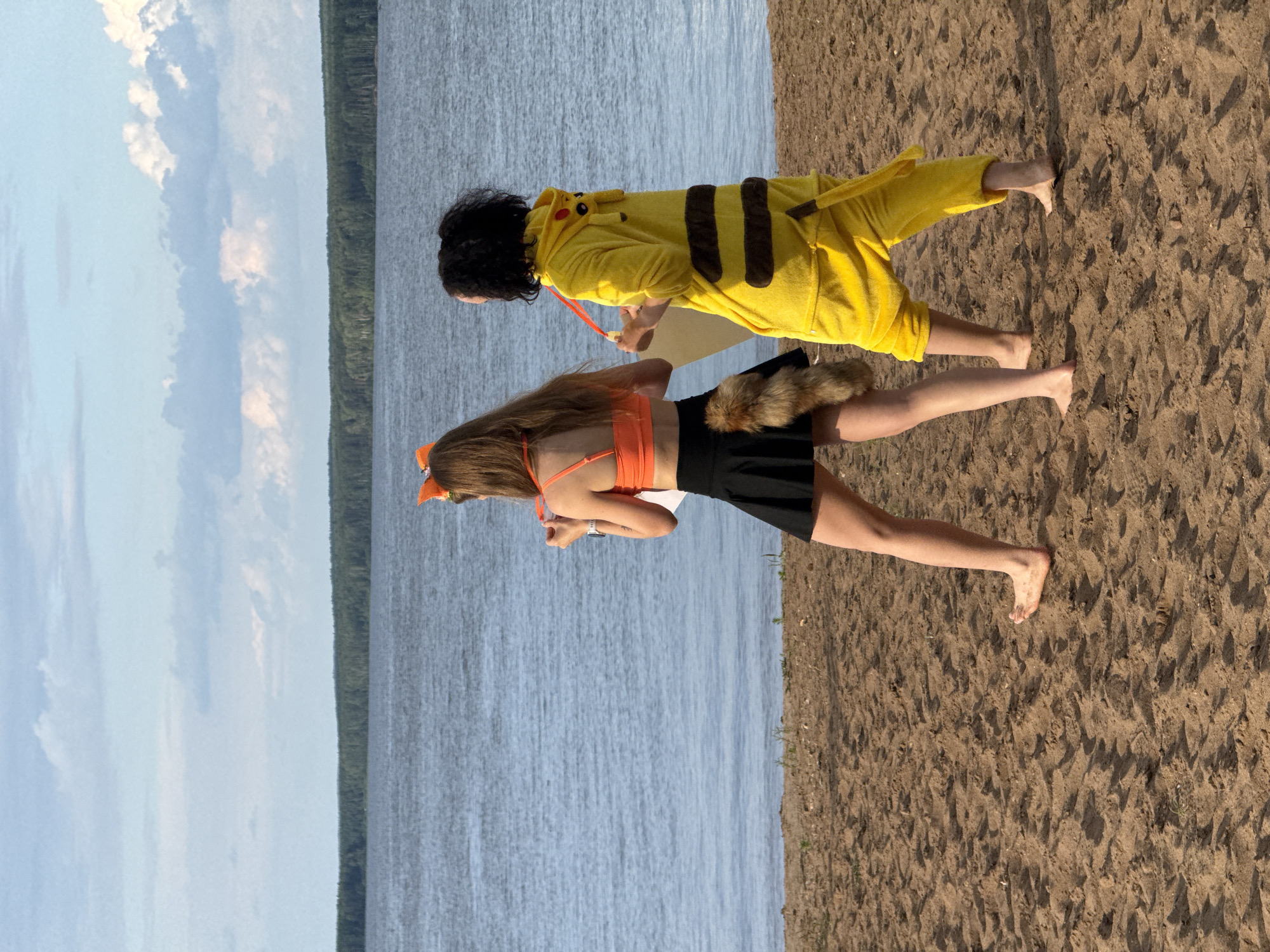 Two individuals, one in a yellow Pikachu costume and the other in an orange top, black skirt, and fox ears and tail, walk on a sandy beach. The Pikachu-costumed individual is on the right, with dark, curly hair and bare feet. The fox-costumed individual is on the left, with long brown hair, also bare feet, and is holding a piece of paper. Both individuals are facing away from the camera. Behind them, a wide body of water stretches to a tree line under a cloudy sky. This scene is set on the shore of the Kama River in Chaikovsky, Russia. The sand is wet and textured, with small pebbles and debris scattered on its surface. The lighting suggests it is late afternoon or early evening.