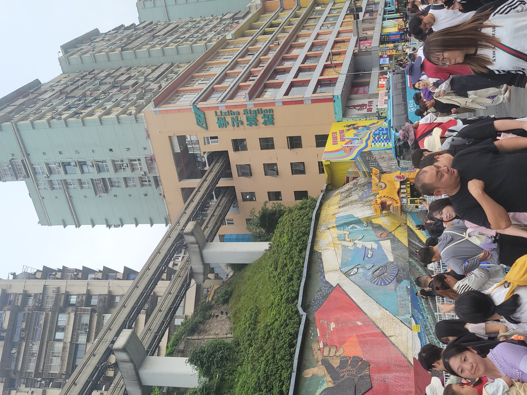 The image depicts a scene in the Yuzhong District of Chongqing, China, showcasing the Liziba Station, renowned for its monorail passing directly through a residential building. A large crowd of people are visible, many seemingly tourists or locals observing or photographing the unique architecture and rail system. The crowd is diverse in age and attire. The station building is prominently featured with the words "Liziba Station" visible on the facade. A section of the monorail track is visible, appearing to pass directly into the building. Adjacent to the crowd is a mosaic wall. There are additional buildings in the background. The weather appears overcast.