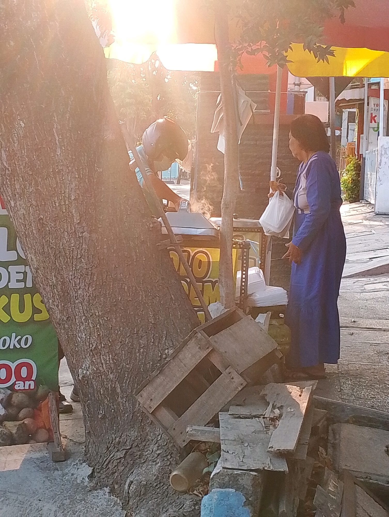 An Indonesian woman in a blue dress stands on a sidewalk in Probolinggo, Indonesia, holding a white plastic bag. She is looking towards a food stall where a person wearing a helmet and mask is preparing food. Steam rises from the food preparation area. The scene is set in the late afternoon, with sunlight casting a warm glow on the surroundings. The sidewalk is made of concrete, and there are wooden crates and a gas tank near the food stall. A large tree with a rough bark trunk is prominently positioned on the left side of the frame, with some text visible on a green sign attached to it. In the background, shops and other structures line the street.