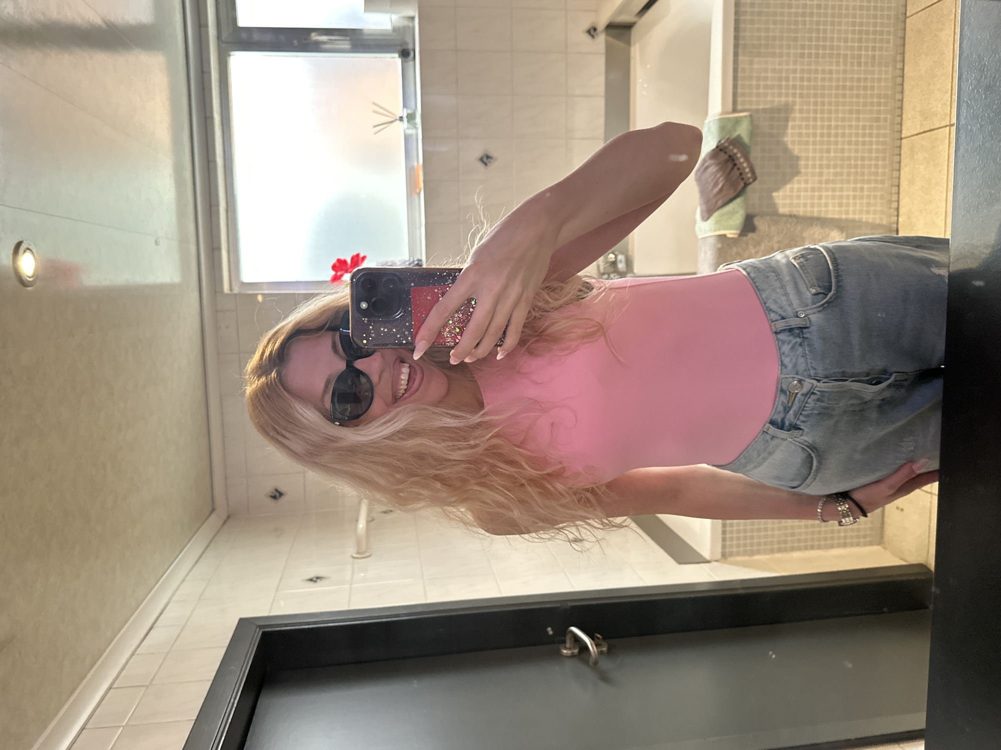 A woman with blonde hair and sunglasses takes a mirror selfie in a bathroom. She is wearing a pink tank top and light blue jeans. The bathroom has tiled walls and a bathtub.