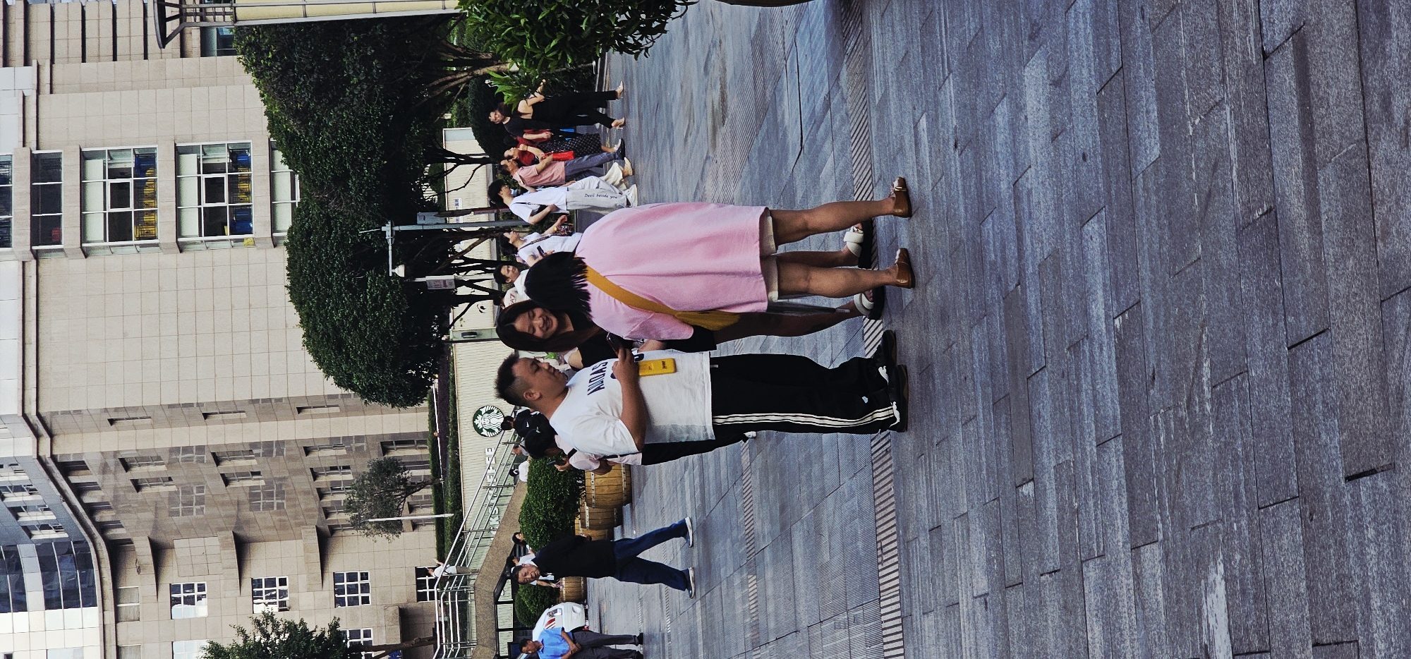 The image shows a group of people standing in front of a Starbucks in Yuzhong District, China. One person wearing a white t-shirt and black pants is speaking to another person wearing a pink shirt. Other people are gathered behind them, seemingly waiting in line. The environment appears to be a paved public area with a building in the background.