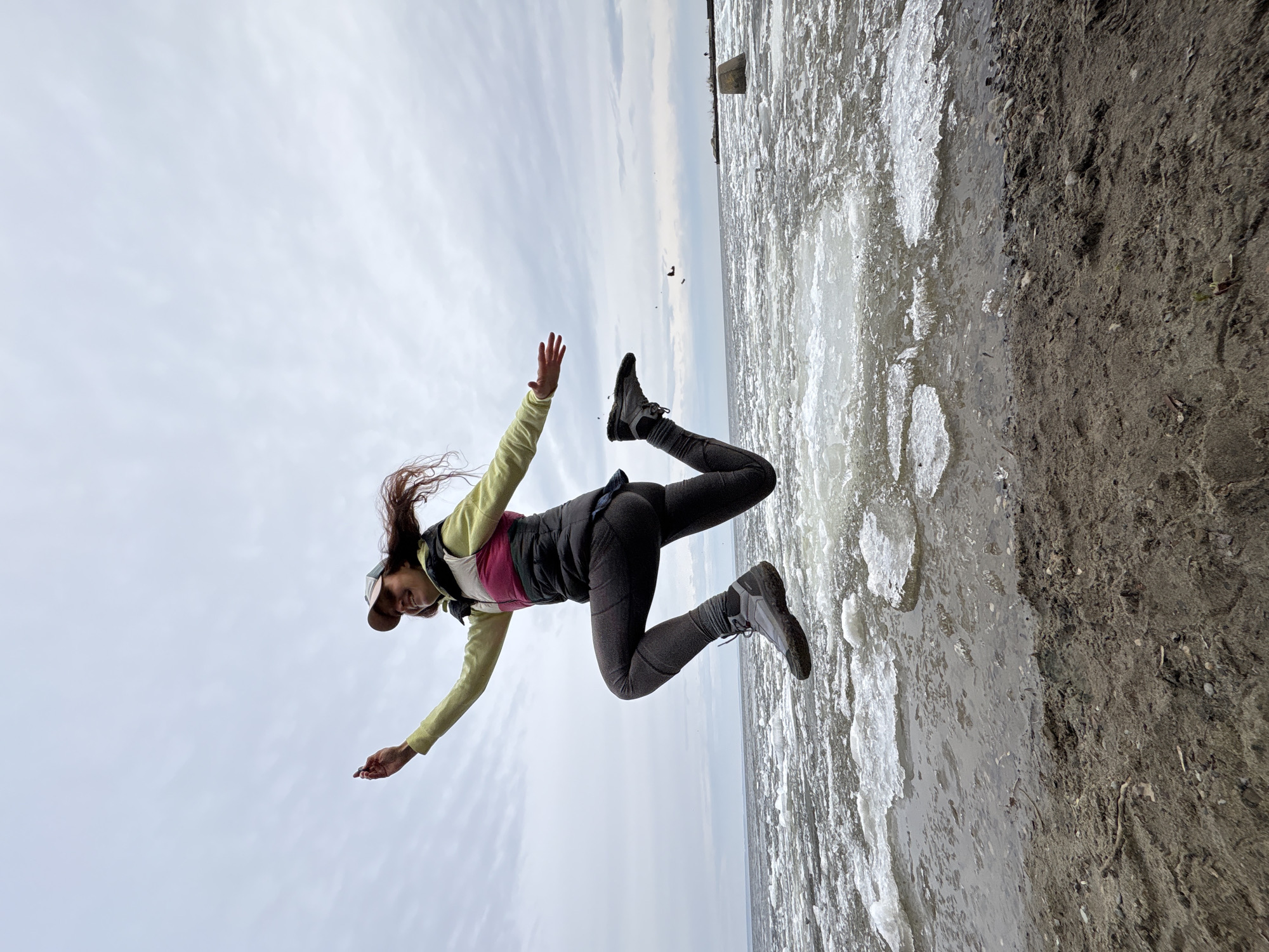 A woman in outdoor clothing is suspended mid-air against a backdrop split between a cloudy sky and a partially frozen, possibly snow-covered or icy water body. She is wearing a cap, a light-colored long-sleeved shirt, a dark vest with a pink accent, dark leggings, and hiking boots. Her arms are extended and her legs are bent. The water appears to be at the edge of a beach or shore with some ice formations. There is a structure visible in the distance over the water. This is in Ulyanovsk, Russia.