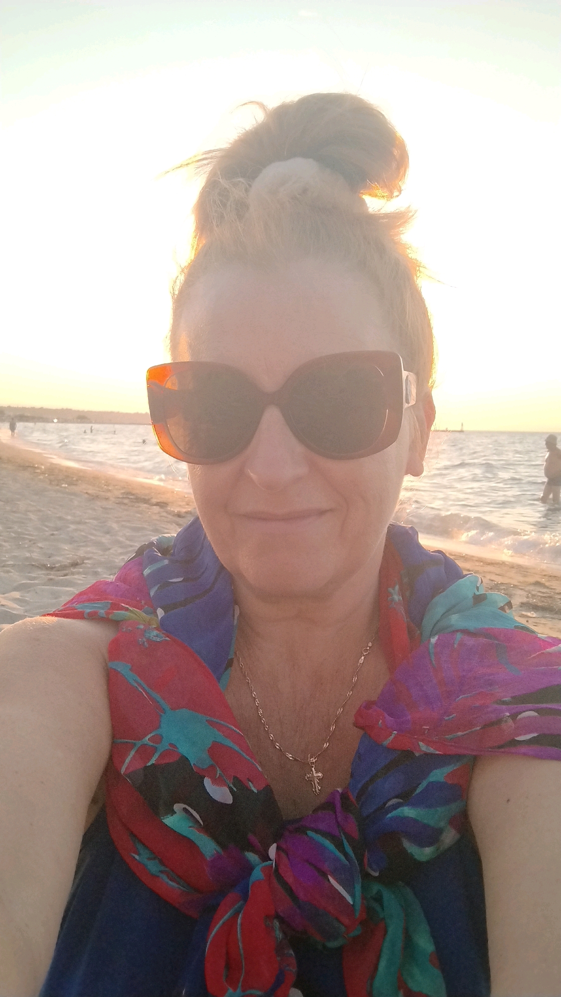 A woman with auburn hair pulled into a bun on top of her head and wearing large, brown-rimmed sunglasses takes a selfie on a beach. She is smiling at the camera and wearing a colorful scarf with abstract patterns of red, blue, teal, and purple tied around her neck. A thin gold chain with a cross pendant is visible around her neck. The background shows a sandy beach with gentle waves lapping at the shore. In the distance, several people are visible in the water and on the beach. The sky is bright with the sun casting a warm glow over the scene, suggesting it is late afternoon or early evening. The location is likely Neoi Epivates, Greece, given the typical scenery of beaches in that area during warmer months.