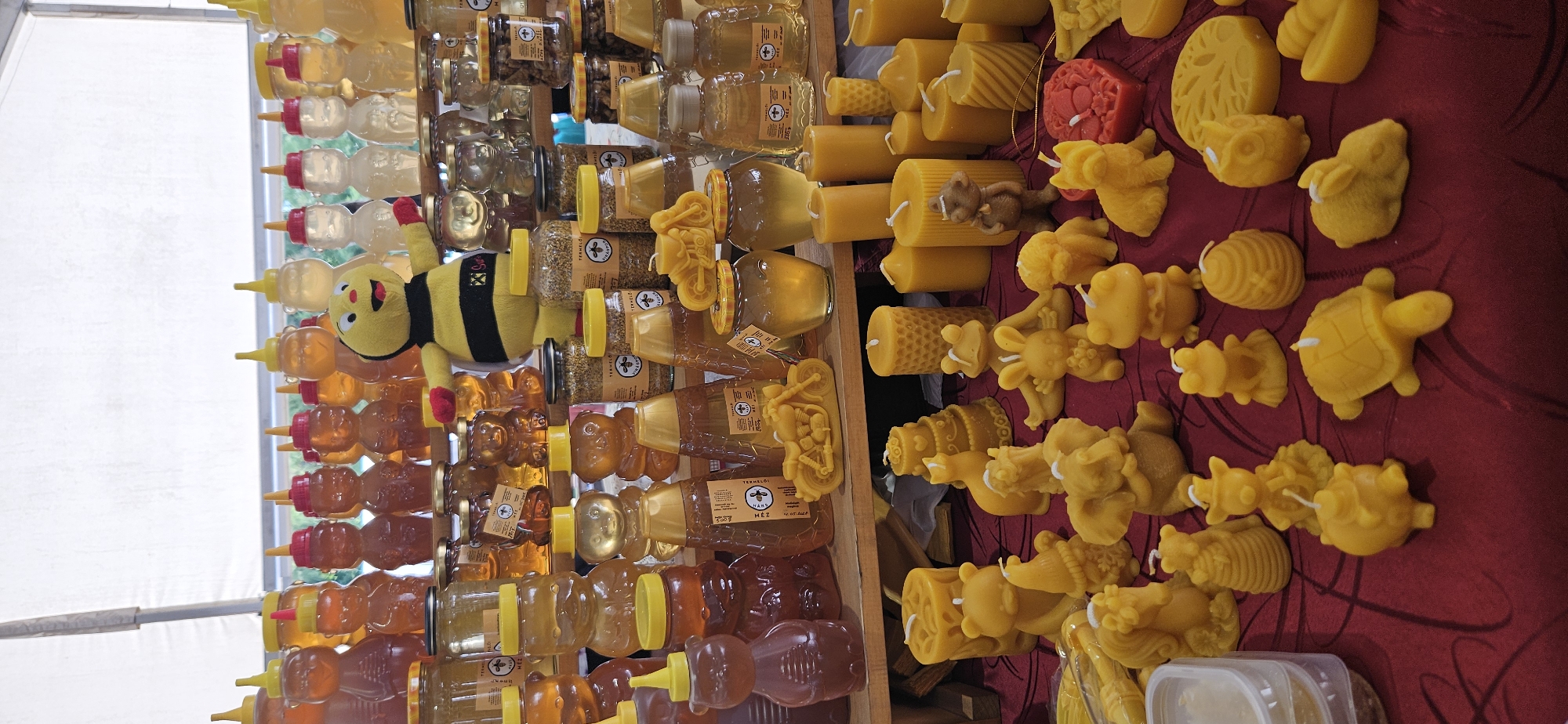 This is a full-frame, eye-level, vertical, medium shot of a market stall in Hévíz, Hungary, displaying a variety of honey products and beeswax candles. The stall is set up on a reddish-brown, patterned tablecloth. In the background, multiple shelves are filled with jars and bottles of honey, many of which are shaped like bears and beehives, with yellow lids. A stuffed bee toy wearing a black and yellow striped outfit is placed prominently on the top shelf, facing the camera.
The foreground is crowded with various beeswax candles. These candles are molded into different shapes, including animals like bears, rabbits, frogs, elephants, dogs, turtles, owls, and squirrels, as well as other objects like a motorcycle, a beehive, a cake, and a tree. Some of the candles are cylindrical or hexagonal pillars. The overall color palette is dominated by the golden hues of honey and the yellow of beeswax, with a few red accents on the bee toy. The lighting appears to be natural, suggesting an outdoor or well-lit indoor market setting. There are no visible people actively interacting with the stall in this frame, though the arrangement suggests a retail environment.