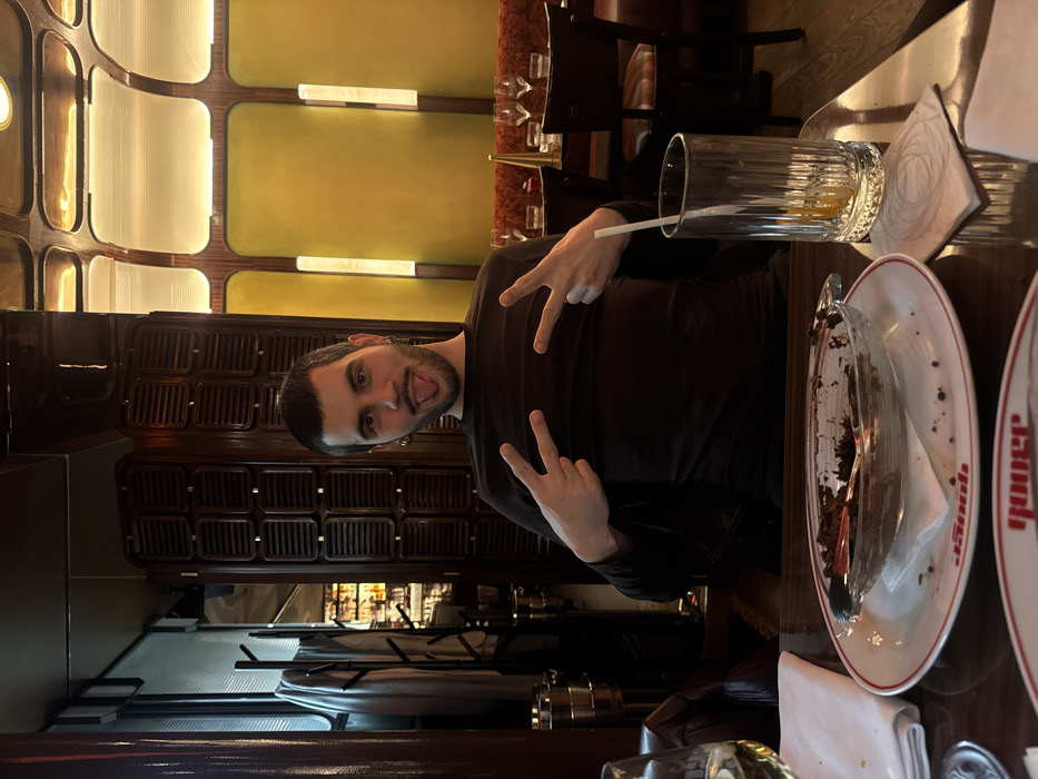 A person poses in a restaurant with a "peace" sign gesture and tongue sticking out. The setting features elaborate wall panels, a bar area, and tables with plates and glasses. One plate has remnants of a chocolate dessert. The restaurant has warm lighting, creating a cozy atmosphere. The person appears to be enjoying their time.