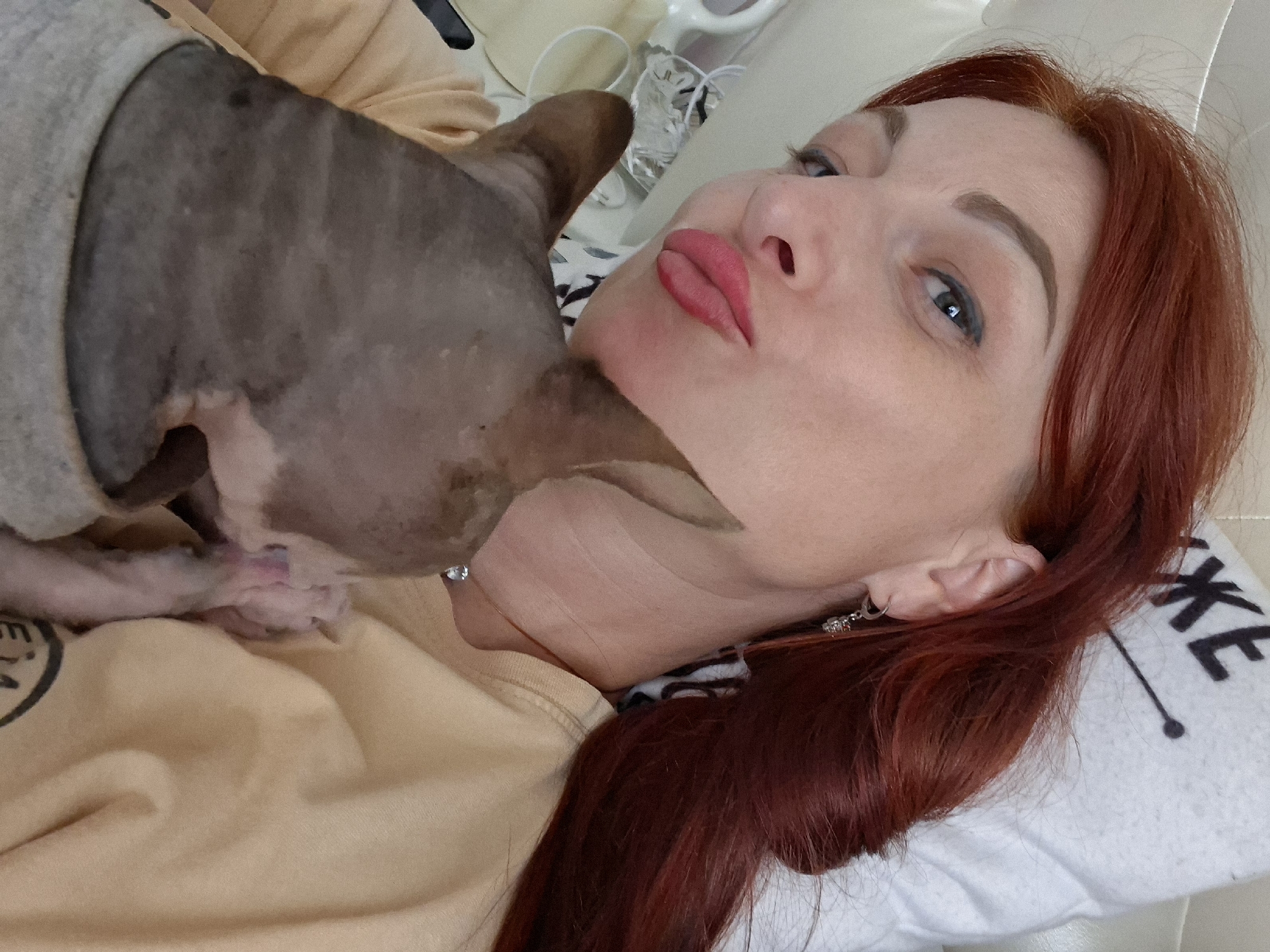 A close-up, angled selfie shows a woman with auburn hair and striking red lipstick lying on a white pillow with black lettering that reads "JK.E". She is holding a hairless cat, likely a Sphynx, with grey skin and pink ears. The cat is nestled into the woman's shoulder. The woman is wearing a tan t-shirt, and a thin silver necklace with a diamond pendant is visible. Her left ear sports a dangling silver earring with a clear stone. The background is softly blurred, suggesting an indoor setting. No specific landmarks of Myadzyel, Belarus are identifiable in this image.