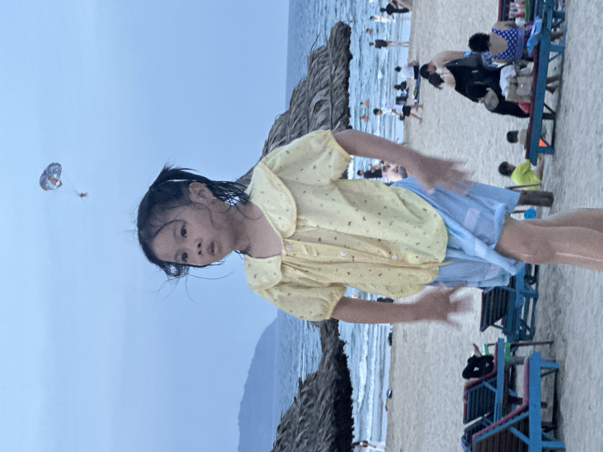 This media file captures a moment at a beach in Da Nang, Vietnam. The primary subject is a young girl in the foreground, wearing a yellow shirt with small red and white polka dots and a light blue skirt. Her hair is dark and wet, suggesting she may have recently been in the water. She is looking directly at the camera with a slightly surprised or curious expression. Her arms are outstretched, as if reaching out or reacting to something.
In the background, the scene unfolds with other beachgoers. The ocean is visible, with gentle waves. A large, thatched umbrella or shade structure stands prominently on the sand. Further in the background, several people are standing in the water or on the sand, some appearing to be playing or relaxing. There are also a few blue beach chairs, some occupied. In the upper left portion of the sky, a small paraglider or kite with an orange and gray design is visible.
The overall lighting suggests it might be late afternoon or early evening, with a soft, diffused light. The sky is a pale blue, indicating calm weather. The presence of beach chairs, umbrellas, and the ocean scene clearly places the activity at a beach resort or public beach. The atmosphere appears relaxed and recreational, with people enjoying a day at the seaside. No visible text is present in the media file.