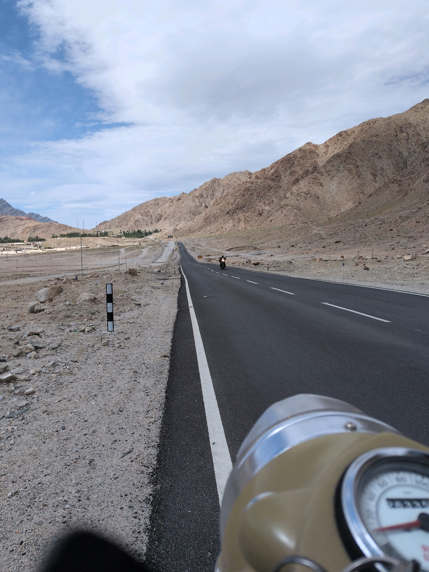 The image depicts a road scene in Upshi, India, with a focus on the road and the surrounding mountainous terrain. A paved road stretches into the distance, marked by a white dashed line and a solid white line on the shoulder. The road surface appears to be in good condition. On the left side of the road, there is a gravelly shoulder with scattered rocks and a few small, sparse green plants. A black and white striped marker post is visible on the left shoulder. In the middle distance, a few buildings and some sparse vegetation are scattered along the base of the mountains. A single motorcycle is seen riding on the road in the distance. The background is dominated by barren, rocky mountains with a light brown hue. The sky above is partly cloudy, with areas of blue sky interspersed with white clouds. The foreground is partially obscured by a tan and silver object, likely part of a motorcycle, with a gauge visible. The overall impression is a remote, arid landscape with a road leading through it.