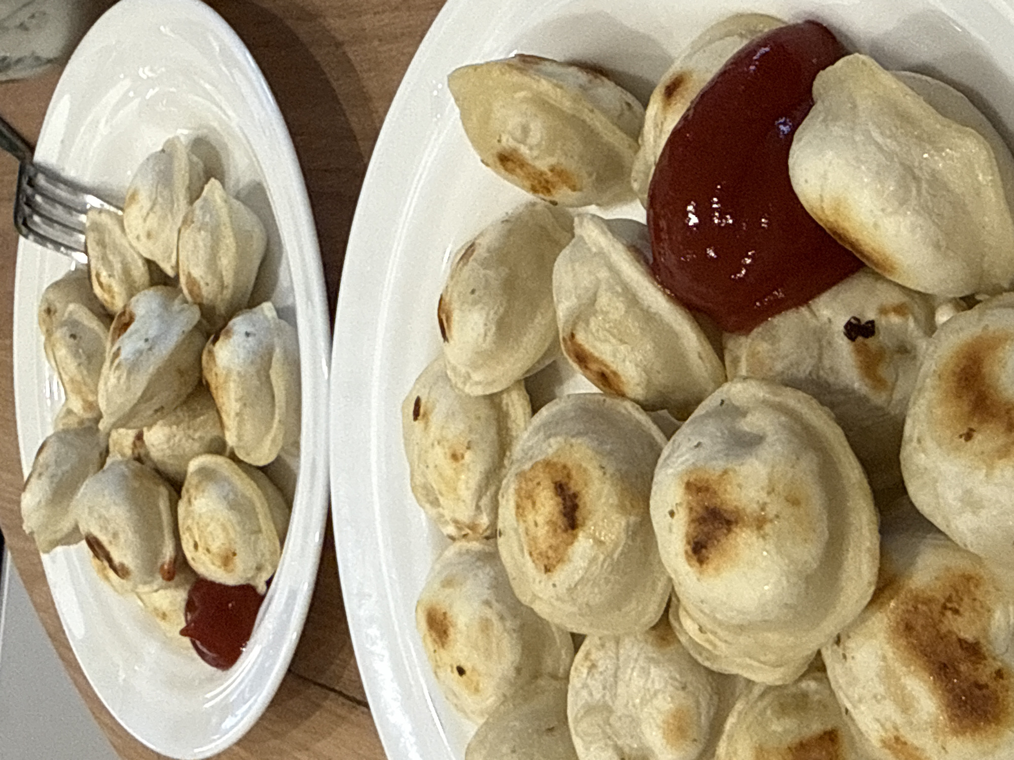 Two white plates of fried pelmeni with ketchup are on a wooden table. A fork is on the plate to the left. The location is Новая Разводная, Russia.