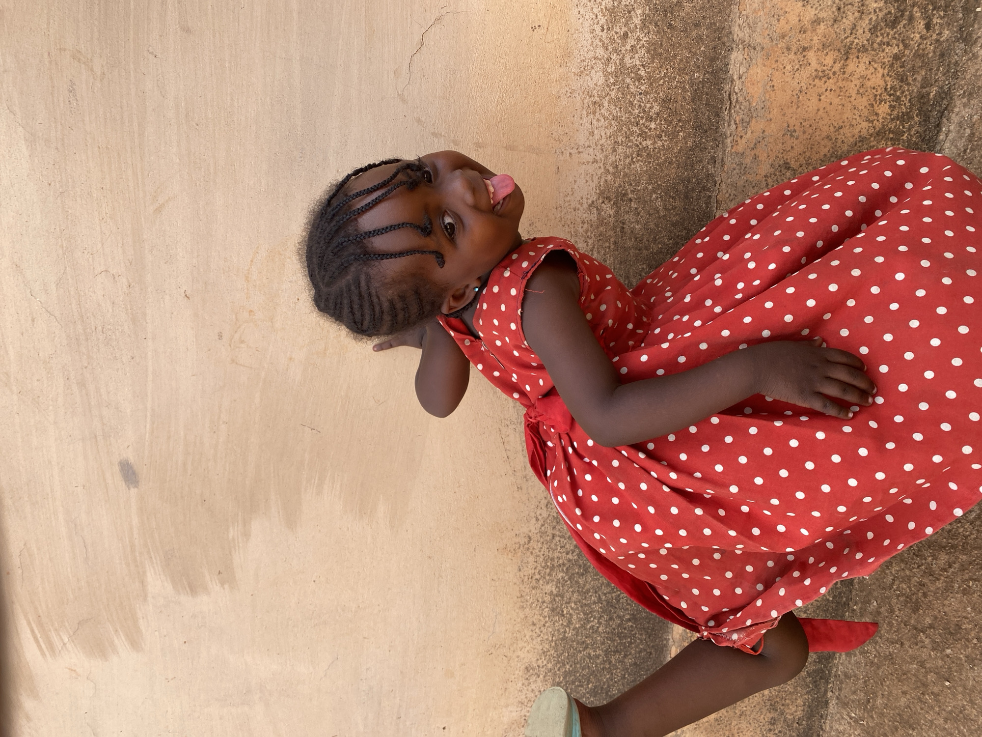 A young child, likely female, with dark skin and braided hair, is depicted in an outdoor setting in Kuru Dungus, Nigeria. She is wearing a vibrant red sleeveless dress adorned with numerous small white polka dots. On her feet, she wears sandals; one appears light blue or green, and the other is red, matching her dress.
The child is the primary subject, positioned slightly off-center and angled. She is looking directly at the viewer with wide, bright eyes and a playful expression, sticking her tongue out. Her arms are bent, resting against her body, and she appears to be standing with a slight bend in her knees, as if posing.
The background consists of a light beige or sandy-colored wall or vertical surface behind her, exhibiting some textured unevenness, cracks, and subtle discolored streaks. Below the wall, a darker, rougher ground surface is visible, suggesting unpaved or textured ground. The lighting is bright and natural, indicative of daytime. No visible text is present in the image. The child's demeanor conveys a sense of happiness and playful innocence.
