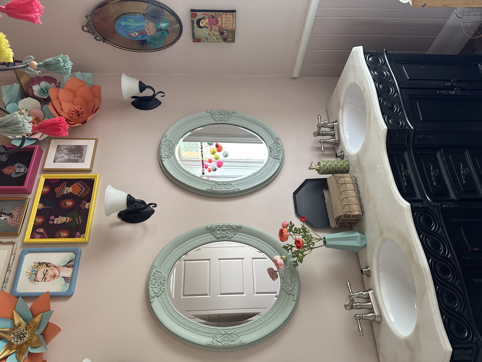 The image depicts a well-decorated bathroom with a pink wall. A double vanity with black cabinetry and a marble countertop is the central feature. Two oval mirrors with ornate light blue frames hang above the sinks. The left mirror reflects a white paneled door, while the right mirror reflects a string of colorful pom-poms and a window.
On the countertop, there are two silver faucets, a green soap dispenser, a teal vase with artificial flowers, and a woven basket holding napkins. Above the vanity, the wall is adorned with various framed art pieces, including a portrait of Frida Kahlo, a yellow framed print with multiple Frida Kahlo-inspired images, and a framed vintage photograph. A large paper flower decoration with orange and teal petals and a gold center is mounted on the upper left wall, along with a cluster of colorful tassels hanging from a chandelier-like fixture above.
On the upper right wall, an oval decorative mirror with a rustic frame contains a depiction of a desert scene with a cactus. Below this mirror is a small rectangular artwork of a woman in a pink dress.
The lower right portion of the image shows a section of pink paneled wainscoting on the wall, and a glimpse of a light wood structure that may be part of a cabinet or shelving unit.
The image does not contain any discernible information that would allow for the identification of its location in the city of Gatineau, Canada, nor does it show any people.