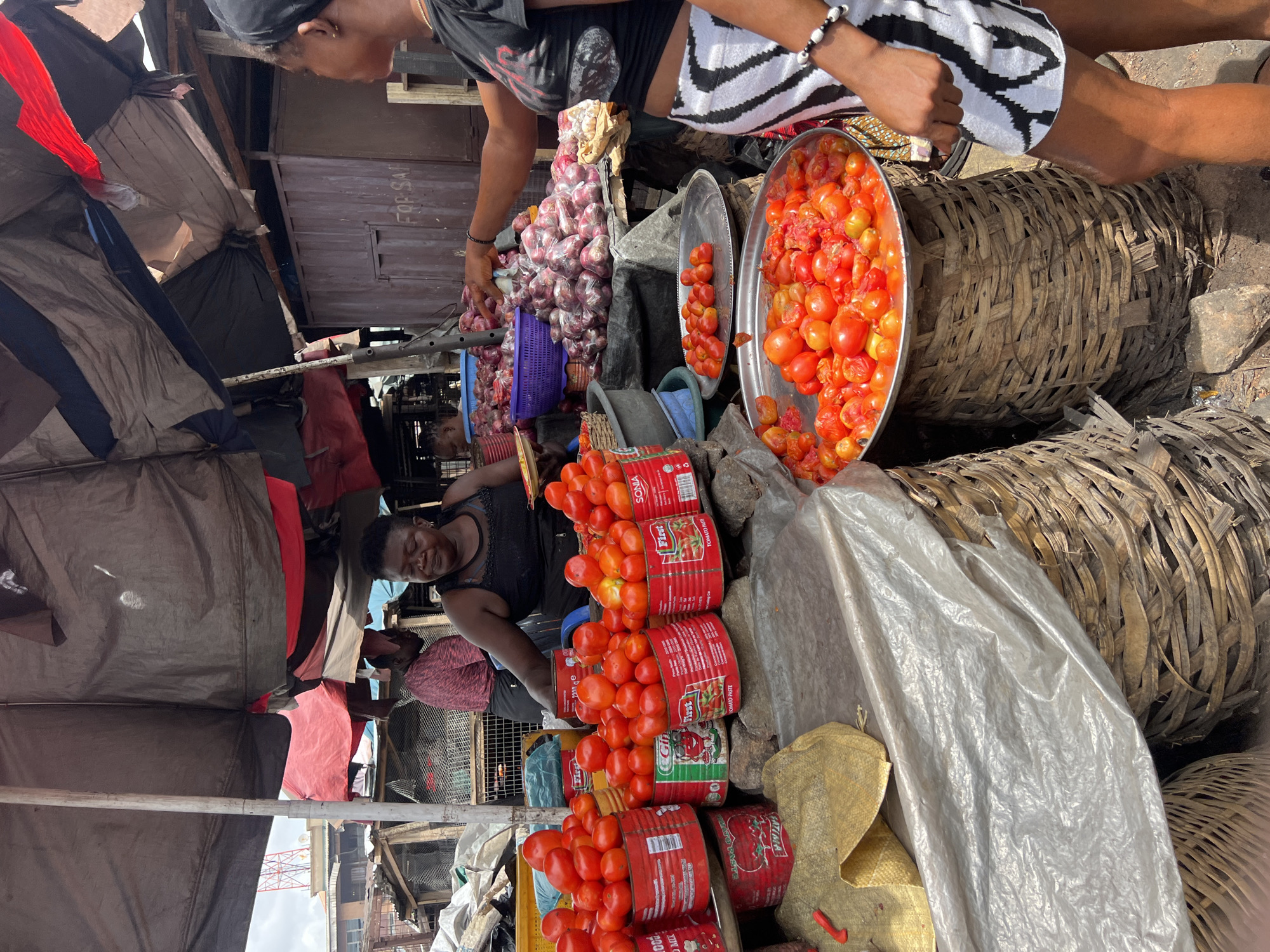 The image captures a bustling scene in Nkpor, Nigeria, likely at a local market. Two women are interacting with a vendor who is seated and selling tomatoes and onions. The vendor is wearing a black top. She sits behind stacks of tomatoes, some arranged on red, cylindrical cans, and is attending to the tomatoes. Above her, another woman wearing a t-shirt with an image on it and zebra-striped shorts is choosing tomatoes from a silver tray. Another pile of onions is in the left corner of the image. To the right, there are two large woven baskets, one of which is partially obscured by a white plastic sheet and a yellow sack. The market appears to be covered by a makeshift roof constructed from various materials, including fabric and plastic sheeting. A building can be seen in the background.