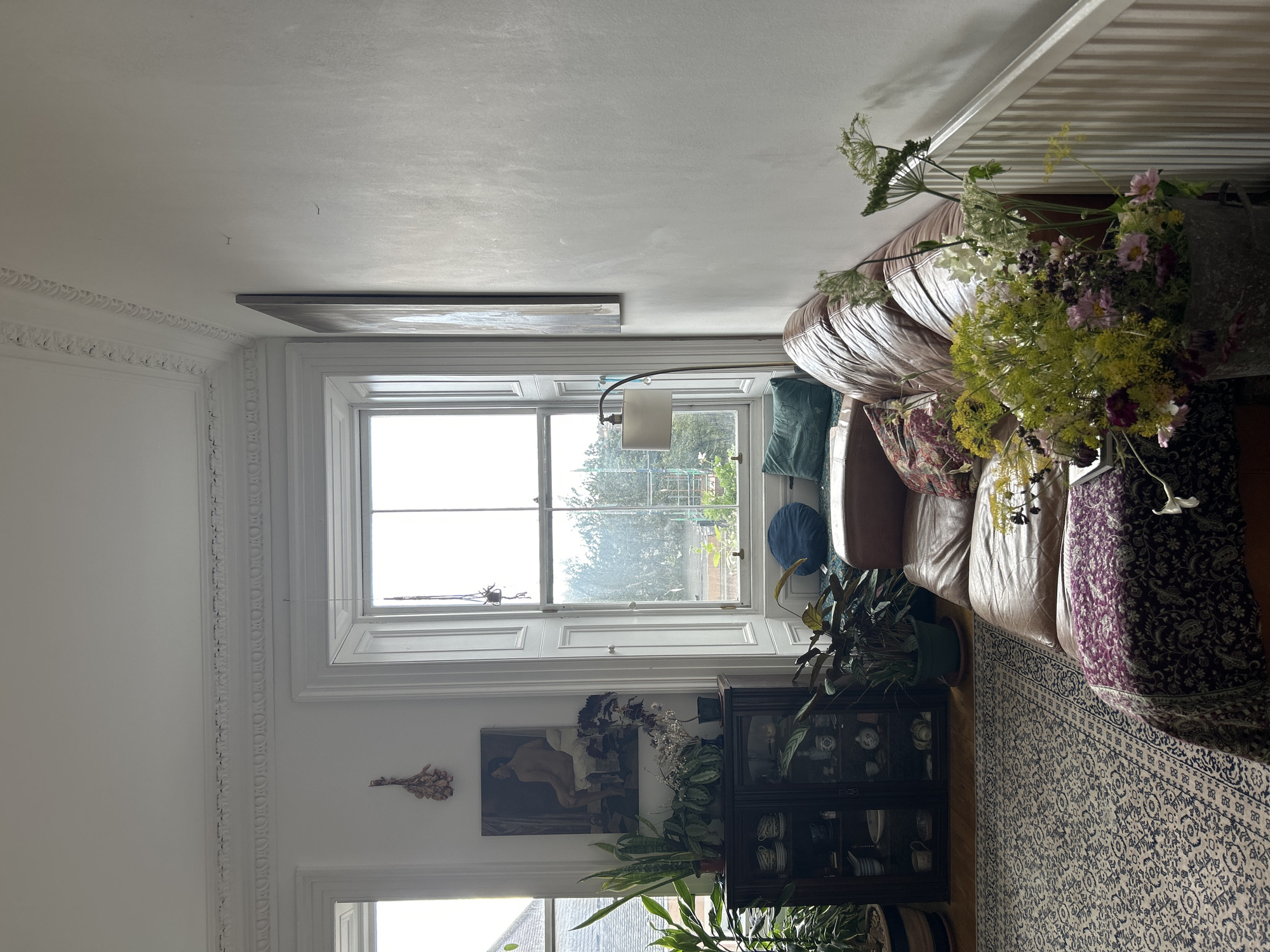The image depicts a sunlit living room in Edinburgh, United Kingdom, featuring a brown leather sofa adorned with colorful cushions and a draped patterned throw. In front of the sofa, a large bouquet of wildflowers is placed in a distressed metal bucket. Behind the sofa, a tall window with white shutters offers a view of greenery and scaffolding. To the left, a dark wooden display cabinet filled with porcelain items is surrounded by numerous potted plants, and a painting hangs on the wall above it. The room has white walls with decorative molding along the ceiling.