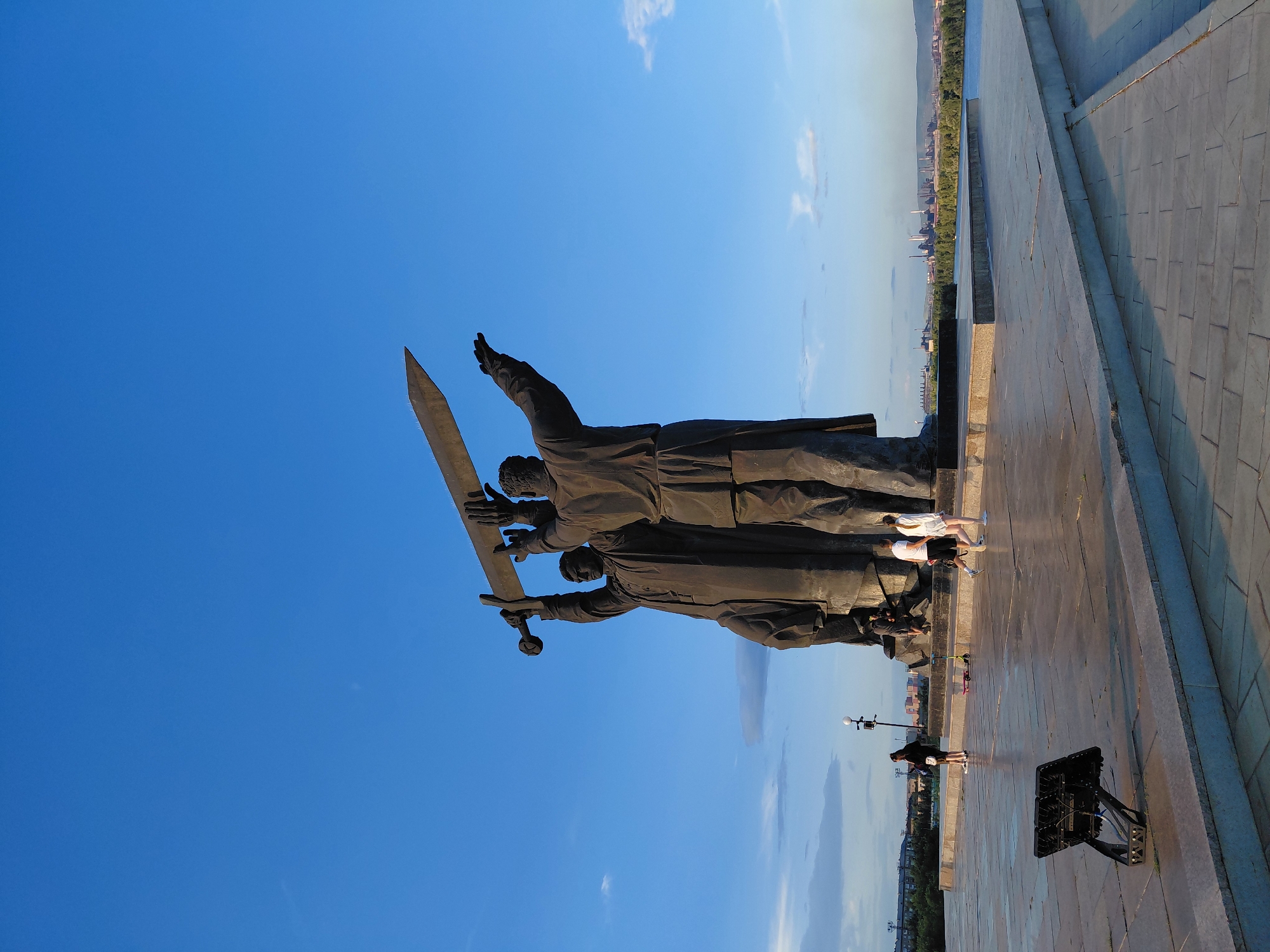 This is a full-frame, eye-level, vertical shot of the Monument to the Magnitogorsk Steelworkers in Magnitogorsk, Russia. The monument, featuring two figures holding a large sword aloft, dominates the center of the frame against a clear blue sky with some wispy clouds. The figures are dark and silhouetted, suggesting it is late afternoon or early evening. In the foreground, a paved plaza stretches out, with a few people walking and standing. A black floodlight is visible on the left side of the plaza. In the middle ground, beyond the plaza, there is a body of water and a distant cityscape with industrial buildings and trees. The overall impression is of a monumental tribute set against a backdrop of urban and industrial life.