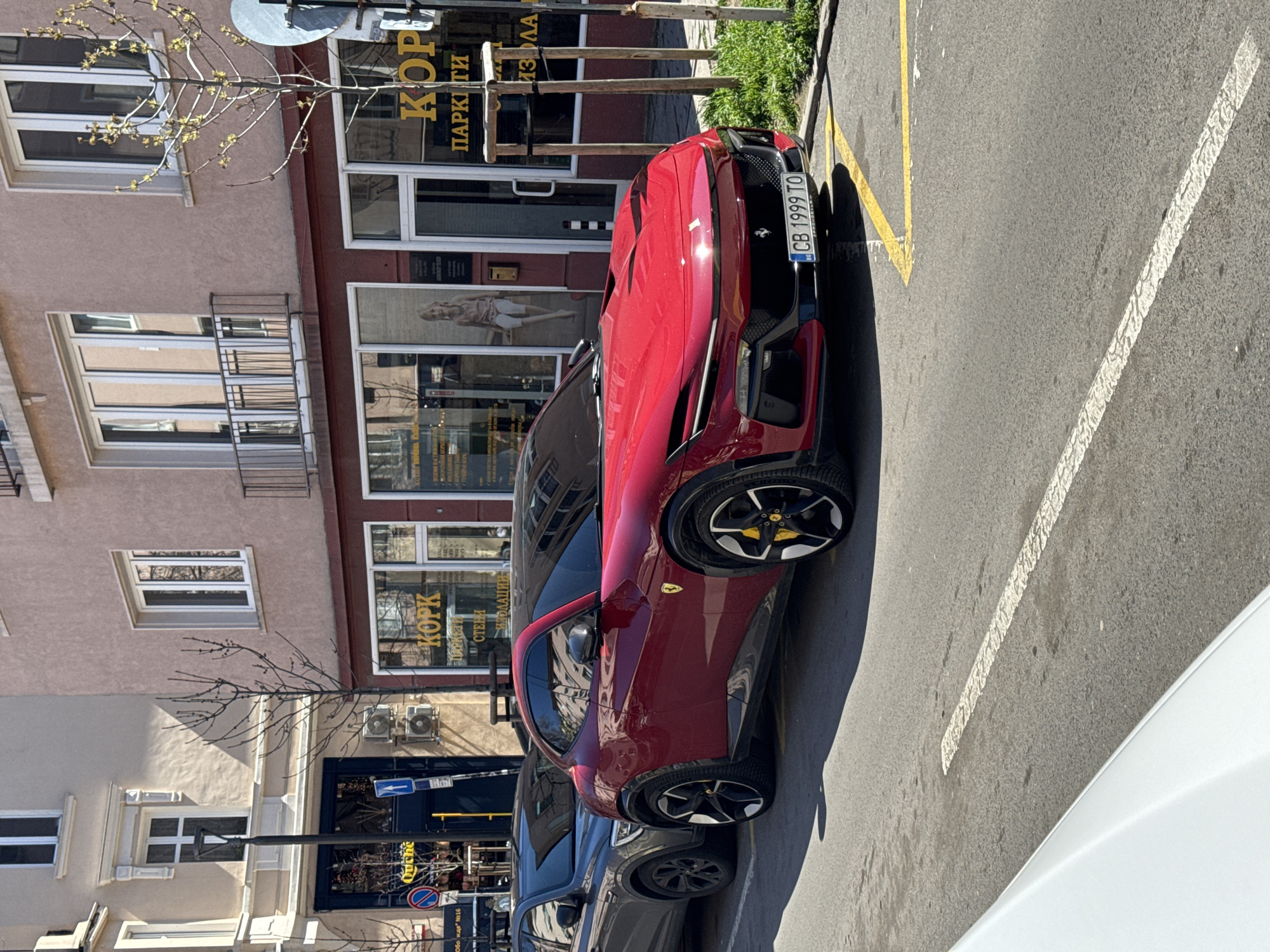 The image shows a red Ferrari parked on a city street in Sofia, Bulgaria. The car is parked in a designated parking spot marked with yellow lines. The license plate of the car is visible, reading "CB 1999 TO". The background features a building with multiple storefronts and windows. One of the businesses has a sign in Cyrillic, mentioning "KOPK" and "ИЗОЛАЦИИ". There is another shop sign with "Ouiche", another shop is "KOPK". There are other cars parked nearby. The weather appears to be sunny.