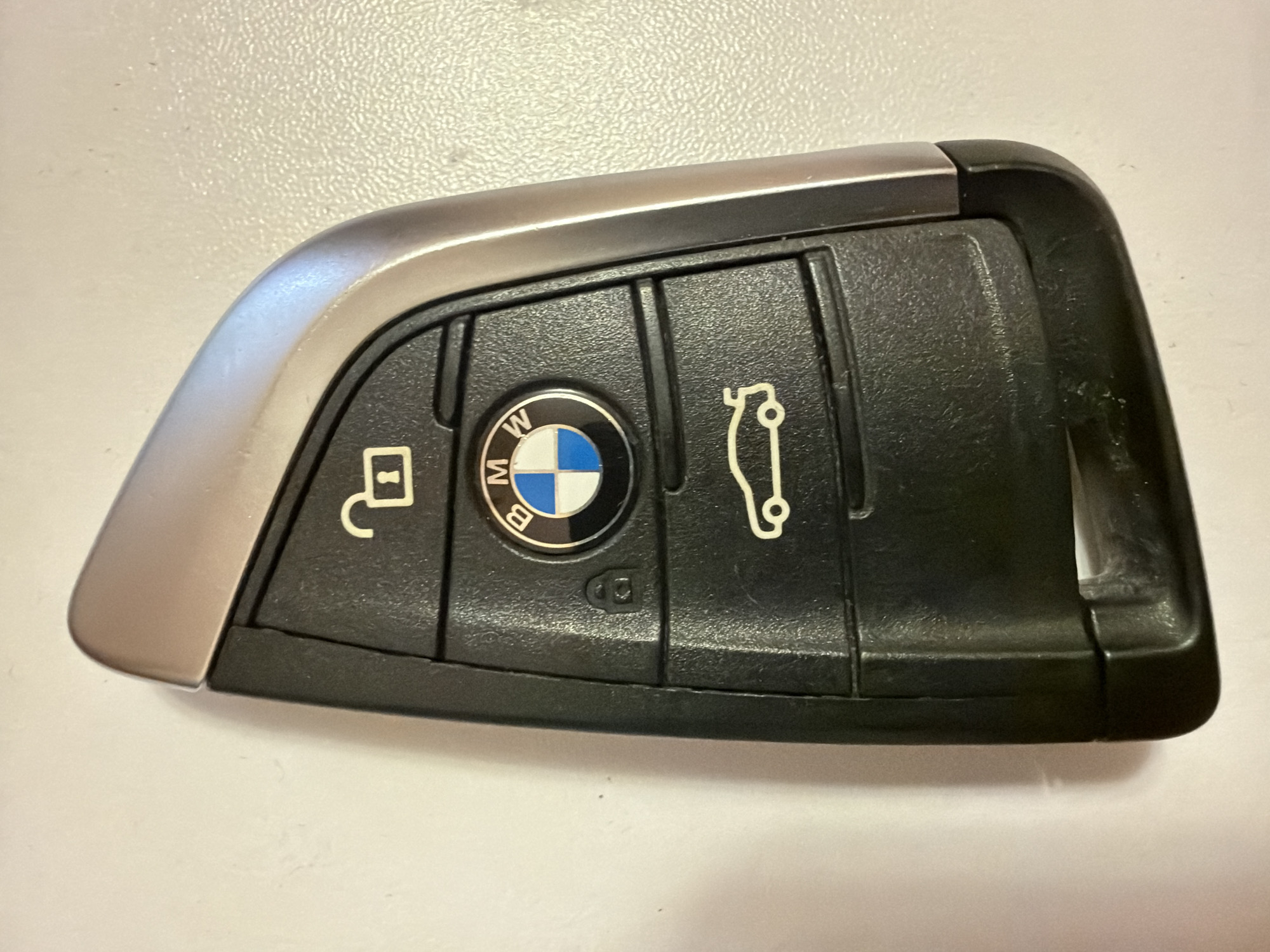 A black BMW car key fob is positioned vertically on a textured, light-colored surface. The key fob has a silver-colored metallic trim along its right edge. The top of the fob features an icon of an unlocked padlock. Below this is the circular BMW logo, with the letters "B", "M", and "W" surrounding the blue and white checkered emblem. Further down, there is an icon of a car with its trunk open. The bottom section of the key fob appears to be a housing for a physical key that is not visible. The lighting creates subtle shadows and highlights on the key fob and the surface. There is no indication of the location in Kyiv, Ukraine, in the image.