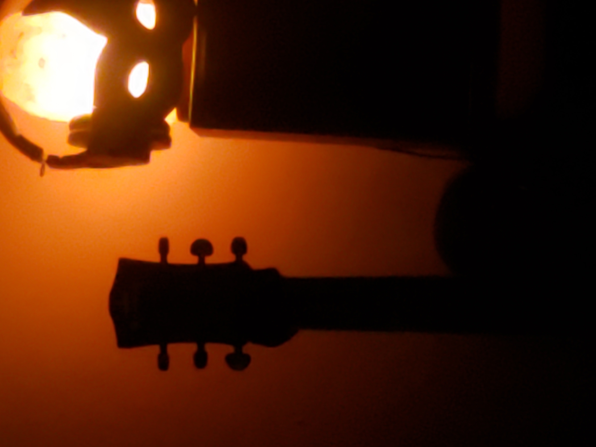 The image shows a silhouette of a guitar neck and headstock on the left, and a silhouette of a lamp with a circular light on the right. The background is a warm orange hue, likely from the lamp. The scene appears to be indoors, with no visible indications of a specific city or location.