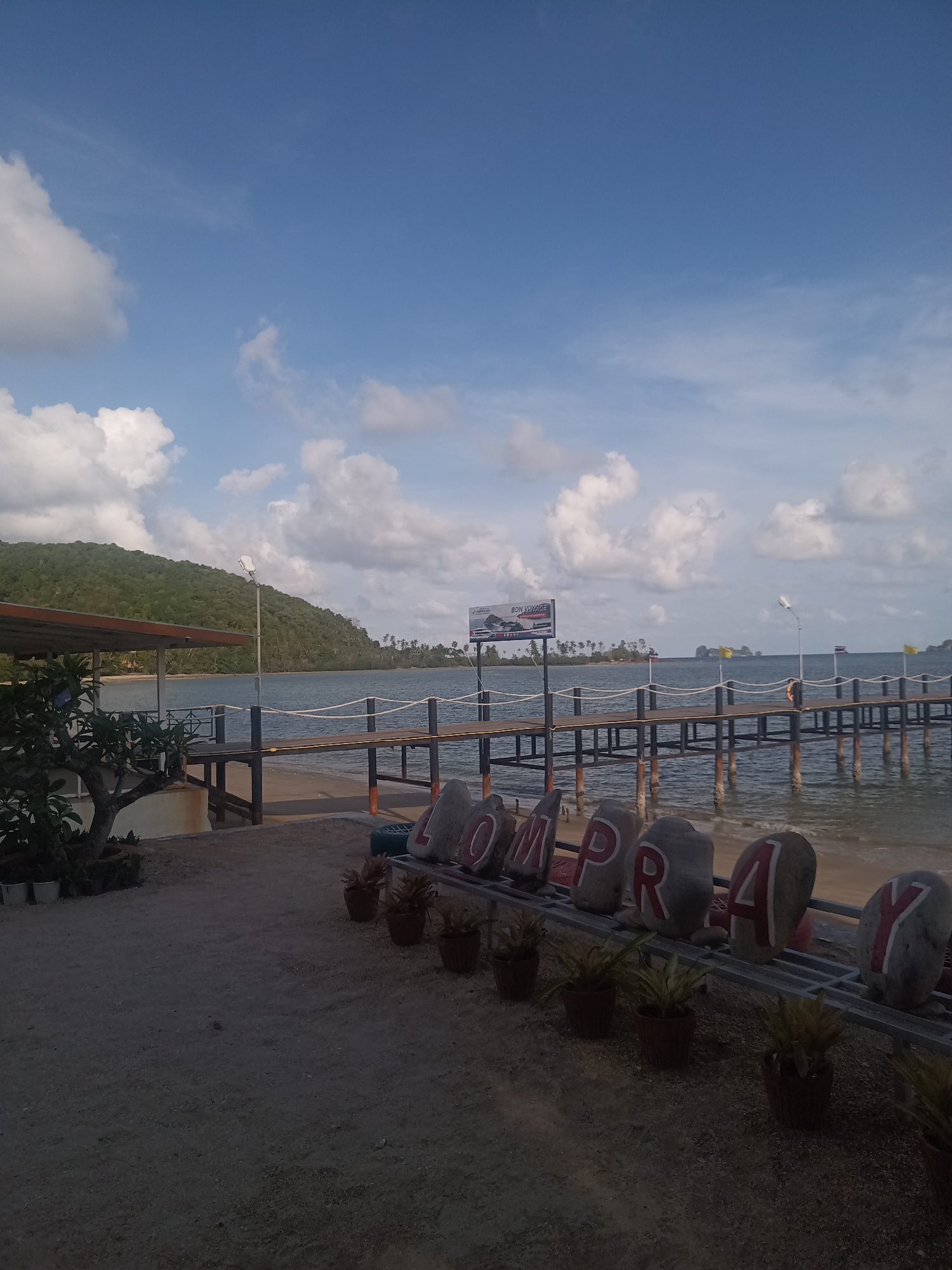 The image depicts a scene at the coast in Tambon Hat Sai Ri, Thailand. A wooden pier stretches out into the water. Along the shore, there is a display of large stones painted with the letters "LOMPRAY", interspersed with potted plants, including bromeliads. The background shows a calm body of water, distant islands, and a partly cloudy sky. A sign with the words "BON VOYAGE" can be seen on the pier.