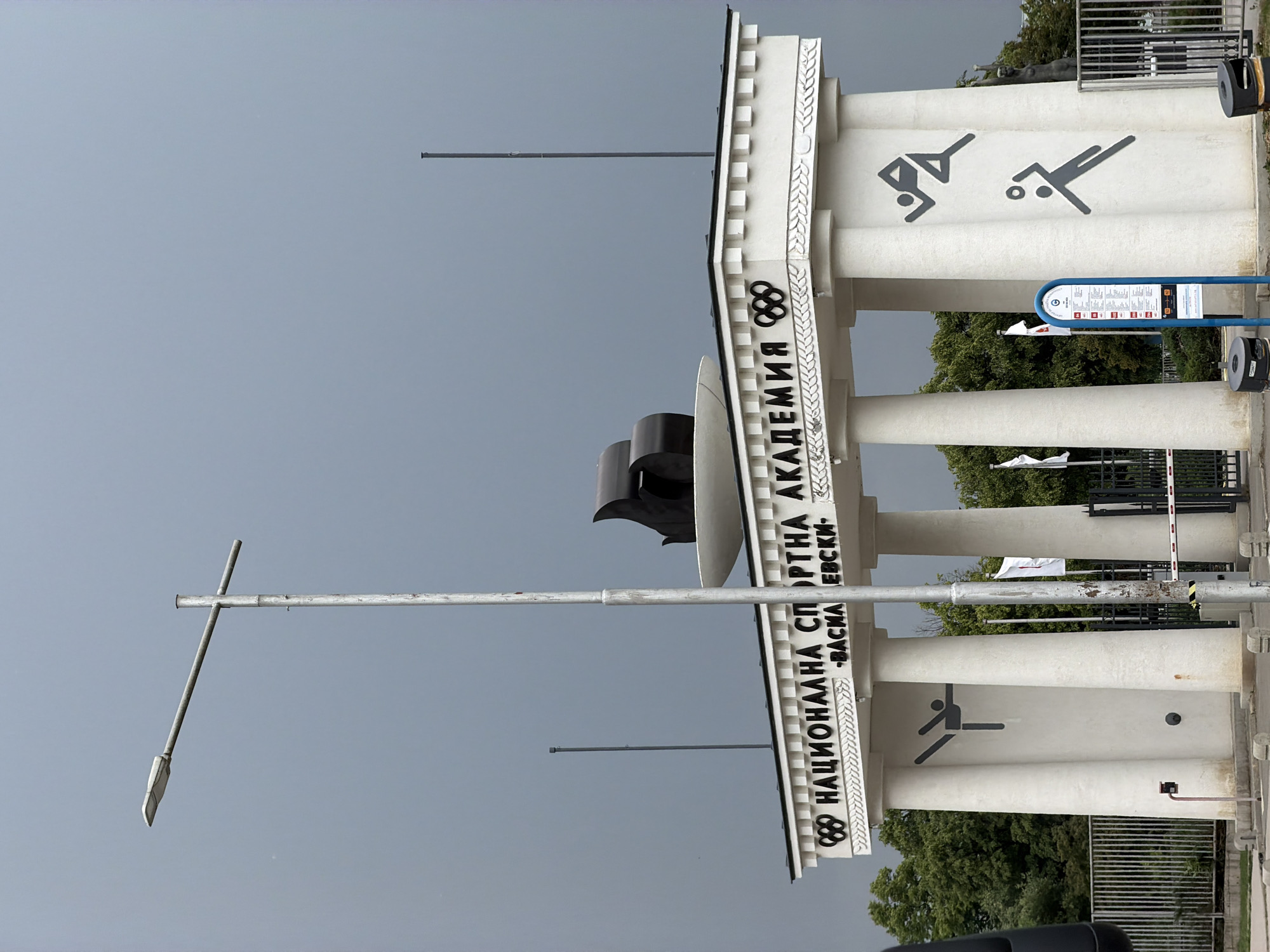 The image depicts the entrance to the National Sports Academy "Vasil Levski" in Sofia, Bulgaria. The entrance is marked by a monumental white structure adorned with the Olympic rings and silhouettes of athletes engaged in various sports. The building features prominent pillars and decorative elements. A streetlamp stands nearby. The sky is overcast.