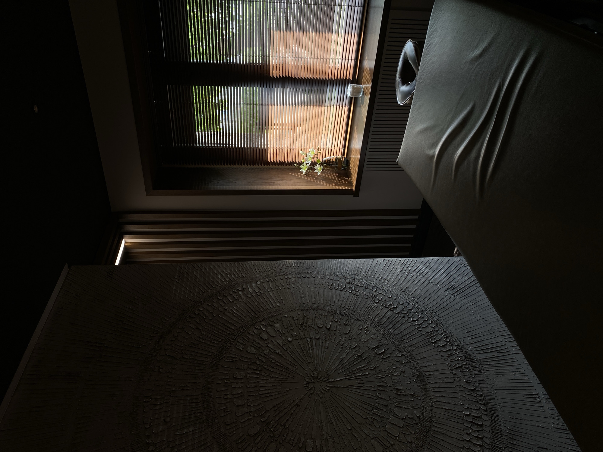 A low-angle, slightly distorted photograph reveals an interior space. A window with slatted blinds occupies the upper center of the frame, letting in natural light. On the window sill, a small vase of flowers sits near a glass container. To the right, a leather chair is partially visible, draped with fabric. Below, a dark, textured painting with a radial pattern dominates the lower portion of the image. The overall scene suggests a dimly lit room with a focus on design elements and natural light. There is no indication that the image was taken in Votkinsk, Russia.