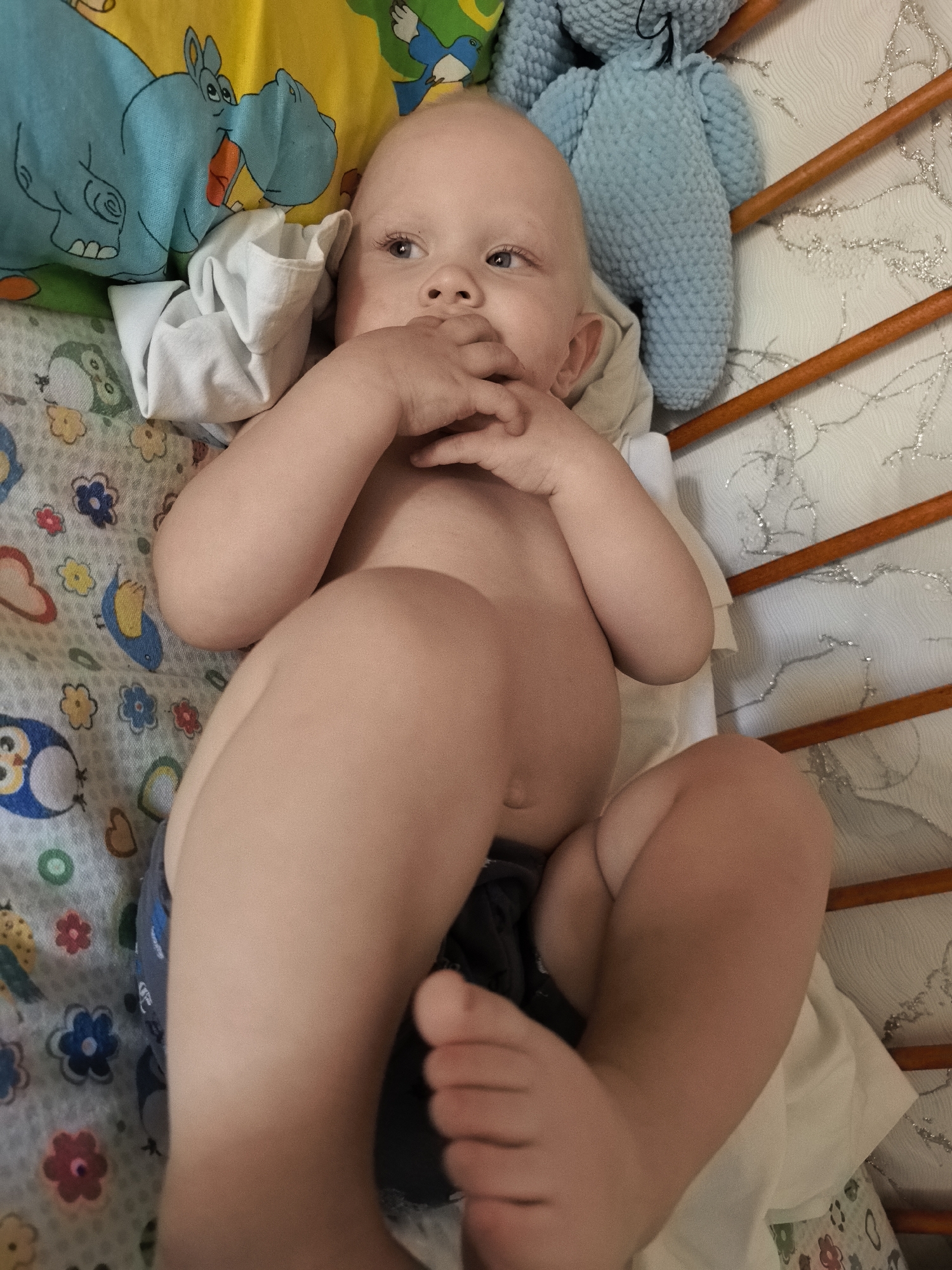 A baby is lying in a crib on its back, with its head turned to the left. The baby is wearing a diaper. There are colorful blankets and a stuffed toy in the crib. The crib has wooden bars. The lighting suggests it is daytime. The baby appears to be a few months old and has its hands near its mouth. The scene is a nursery or bedroom.
