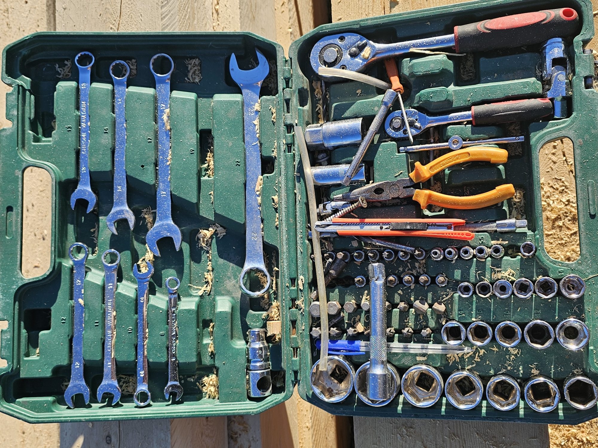 An open, green plastic toolbox filled with various metal tools, including wrenches, sockets, pliers, and a retractable knife, rests on a surface of light-colored wooden planks. Sawdust is scattered throughout the toolbox and on the surrounding wooden surface. The toolbox is positioned outdoors, suggested by the natural lighting and the wooden planks which appear to be construction material or firewood. The scene does not provide any specific information to link it to the city of Mayya, Russia, or any particular urban or rural setting. There are no people or actions depicted in the image.