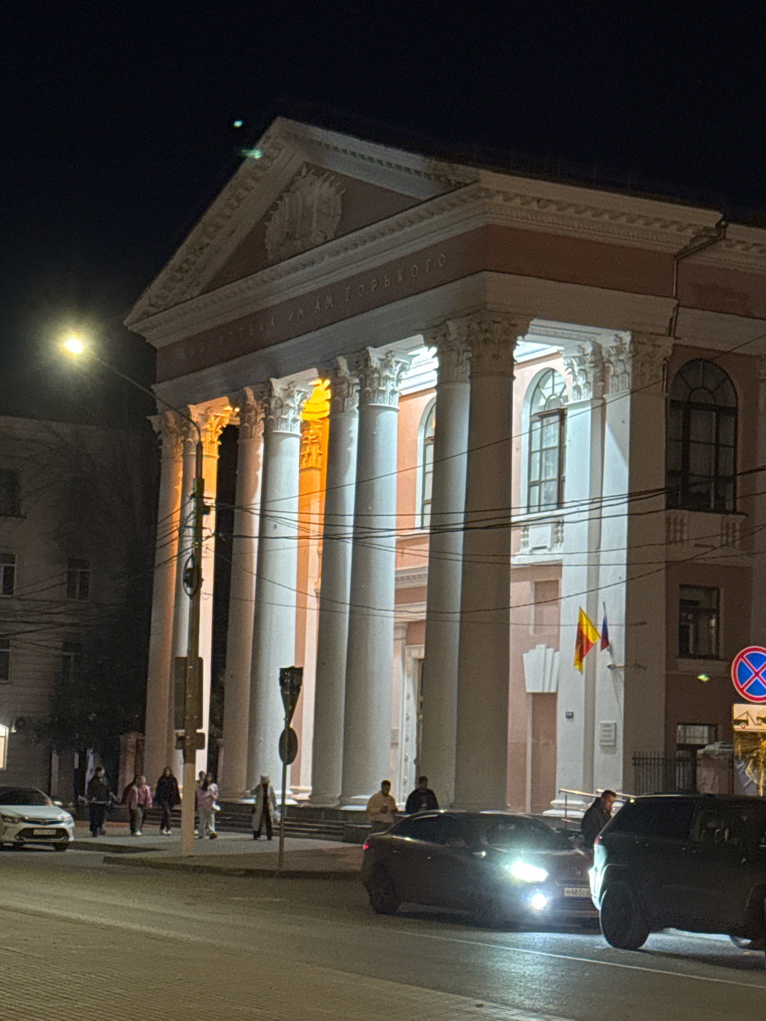 The image captures a nighttime view of the Tver State University building, identified by the inscription "ТВЕРСКОГО ИМ. М. ГОРЬКОГО" (Tver State M. Gorky) on its facade. The grand neoclassical architecture is illuminated by artificial lights, highlighting its imposing columns and pediment. A street lamp casts a warm glow on the left side of the frame.
In the foreground, a street is visible with two cars, a dark SUV on the right and a dark sedan with its headlights on, parked on the left. Several individuals are seen near the building's entrance and on the sidewalk. A group of four people are walking away from the camera near the cars, while another person in a light-colored coat stands on the steps to the building, appearing to gesture. Two flags, one yellow and red and the other blue and white, are mounted on the building. A traffic sign indicating a prohibition for all vehicles is visible on the far right. The scene suggests a late evening or night in a public urban space.