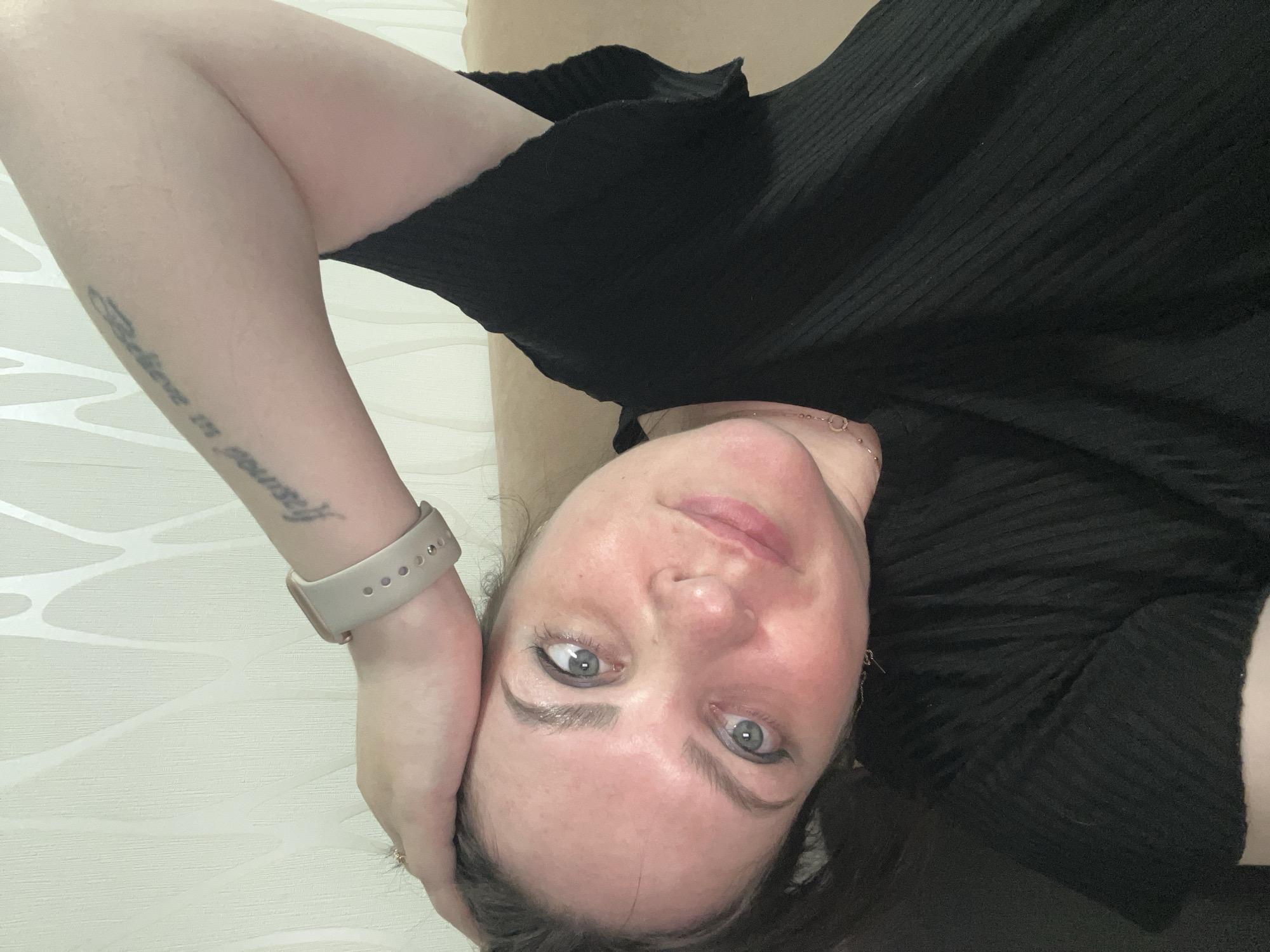 An adult female is depicted in a close-up, slightly tilted portrait shot, lying down and looking directly at the viewer. Her expression is calm and neutral, with a hint of a subtle smile. She has dark hair, fair skin with some natural redness on her cheeks and nose, and striking blue-grey eyes accentuated with subtle eyeliner. She is wearing a black ribbed top or dress. Her left arm is raised, with her hand possibly resting behind her head, revealing a light beige-colored wristband or smartwatch on her wrist. A script tattoo is clearly visible on her left forearm, which reads "Believe in yourself". The background consists of a light-colored wall with a subtle, geometric wave-like pattern, and a beige or light brown surface beneath her, likely a couch or bed. The indoor setting appears well-lit, suggesting daytime. The location is Perm, Russia.
