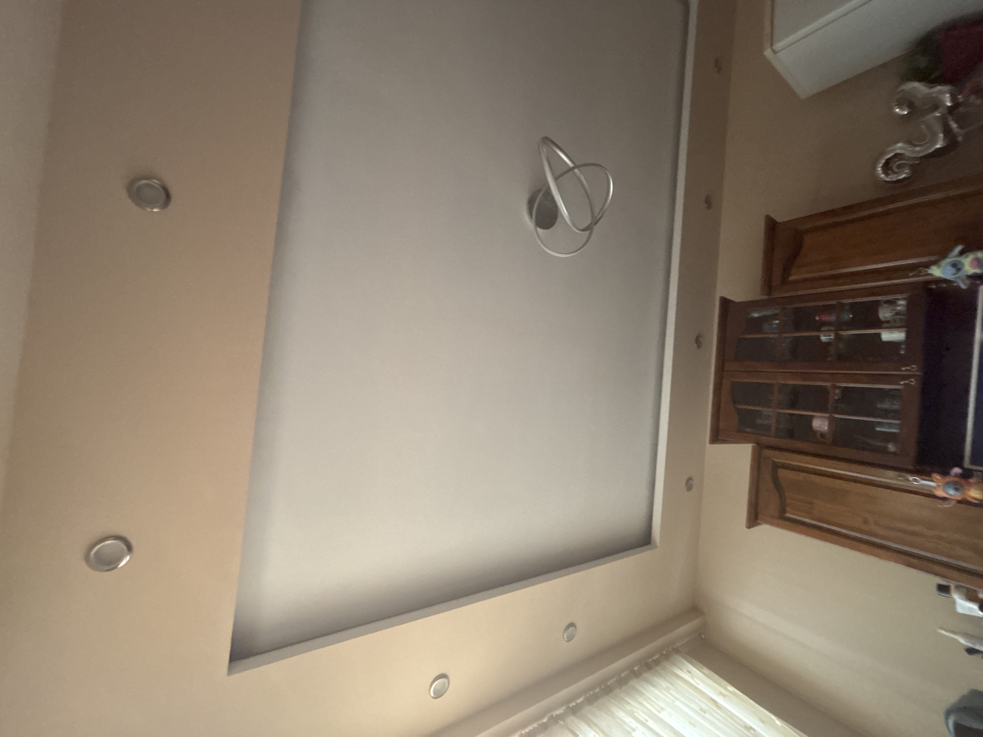The image displays an indoor residential ceiling and the upper portion of a room, purportedly located in Burgas, Bulgaria. The ceiling features a multi-level design: a central, rectangular section painted a light off-white color, surrounded by a perimeter band painted in a beige hue.
Four circular recessed spotlights with silver metallic trim are embedded in the beige perimeter band; two are visible on the left side, one near the top edge, and one on the right. A modern, sculptural light fixture hangs from the center of the lighter ceiling section, characterized by intertwined silver-colored metallic loops.
The visible walls are painted the same beige color as the outer ceiling band. On the far left, a window partially obscured by light-colored, vertical-striped curtains or blinds is visible, indicating a source of natural light.
The lower section of the image shows the top portions of furniture and appliances, suggesting a kitchen or dining area. Prominently featured is a dark wooden cabinet with glass-paned doors, displaying various items such as cups and glasses. A small, colorful plush toy hangs from one of the cabinet handles. To the right of this glass cabinet, a tall, dark wooden cabinet or door panel is visible. On the far right, a section of a white appliance, possibly a refrigerator, and a reflective, ornate silver-colored decorative object are present. No individuals are present in the scene.