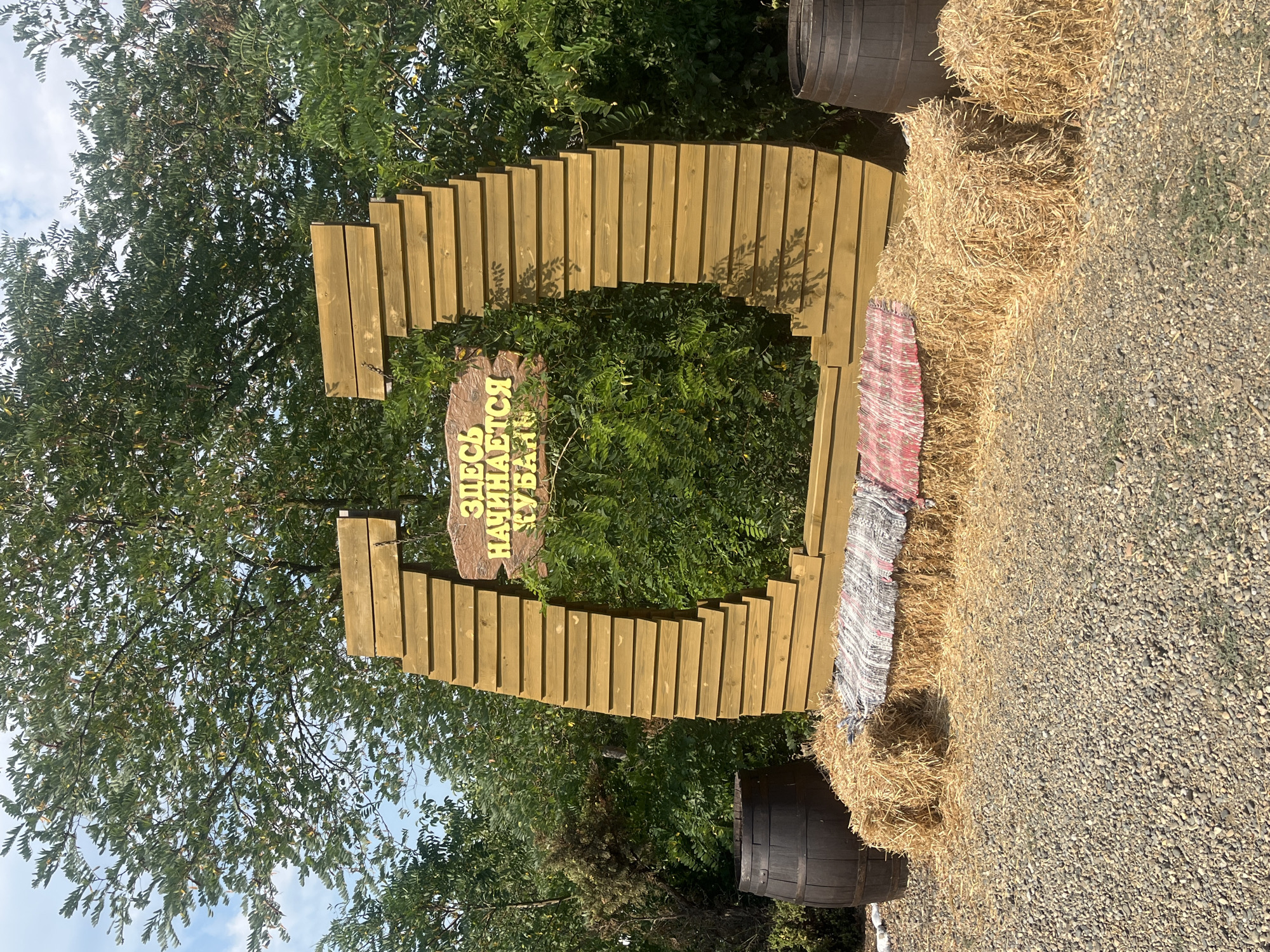In Taman', Russia, a rustic outdoor scene is presented, featuring a large, wooden archway constructed from horizontal planks that are staggered to create a visually dynamic effect. Inscribed on a rustic sign hanging from the archway are the Russian words "ЗДЕСЬ НАЧИНАЕТСЯ КУБАНЬ," translating to "Kuban Begins Here." The arch frames a dense green bush. In front of the archway, several bales of hay are arranged, with one bale serving as a makeshift seat, covered by a colorful striped rug. To the left of the hay bales, a dark wooden barrel stands on a gravel ground. To the right of the hay bales, another similar dark wooden barrel is visible. The background is filled with lush green trees and foliage under a bright sky with some clouds. The ground is covered in grey gravel.