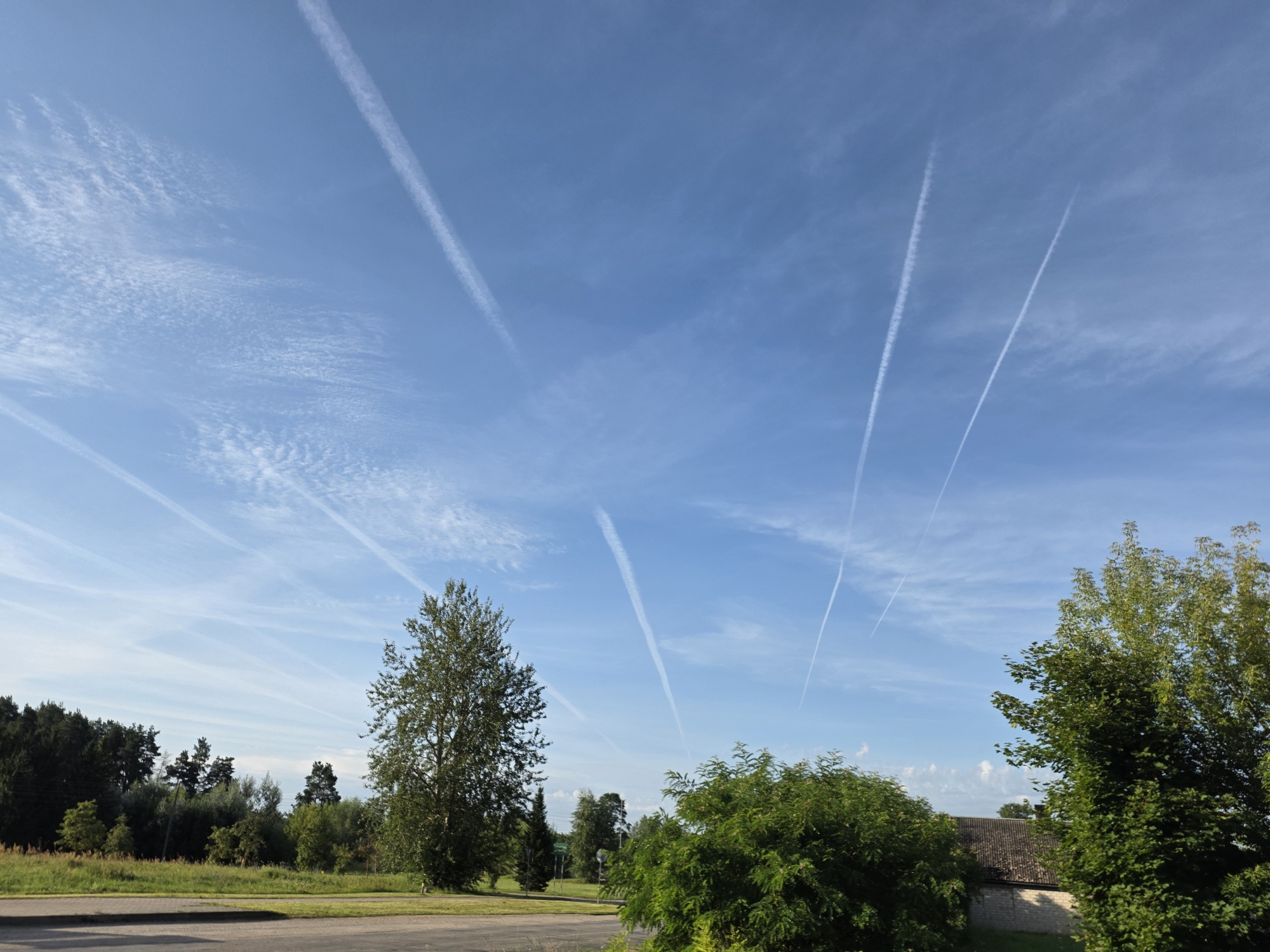 The image shows a clear blue sky with numerous white contrails, indicating airplane activity. Below the sky is a landscape featuring a variety of trees, including deciduous and coniferous varieties, with dense foliage. In the foreground, there is a paved surface, possibly a road or path, and green grass. To the right, part of a building with a tiled roof is visible. The scene suggests an outdoor environment, likely in a rural or semi-rural area, given the prevalence of trees and open sky. The presence of contrails suggests it is daytime. The location in the city of Svēte, Latvia, is not visually ascertainable from the image itself.