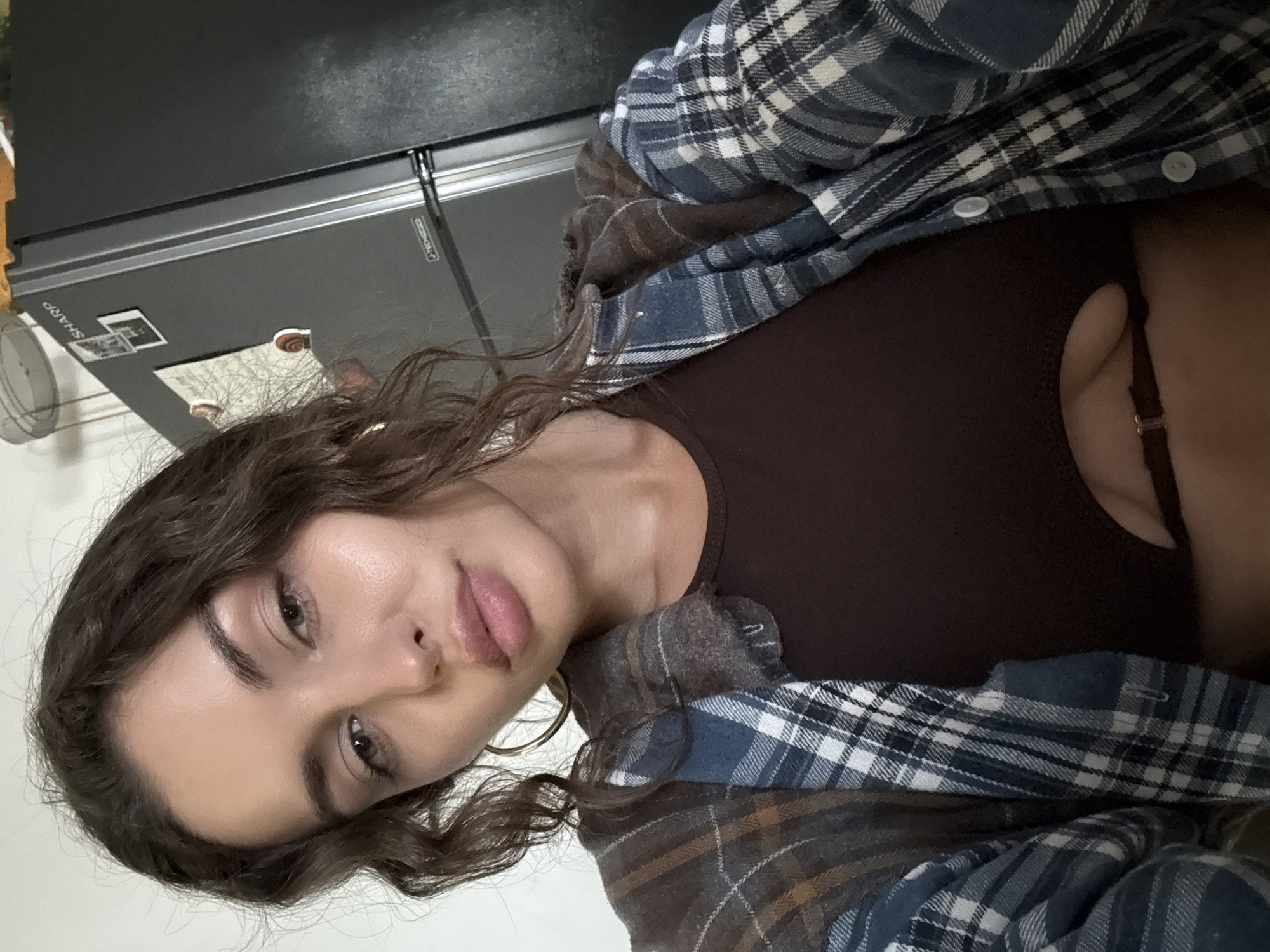 A fair-skinned woman with wavy brown hair is posing for a selfie. She has brown eyes, full lips, and gold hoop earrings. She is wearing a brown top and a blue and brown plaid flannel shirt. The background includes a silver refrigerator with magnets and stickers on it, and a white wall. The location of North Kuta, Indonesia, is not discernible from the image.