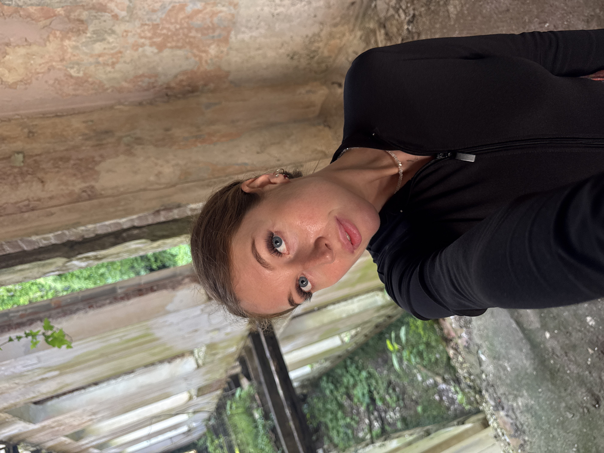 A woman with fair skin, blue eyes, and brown hair is posing for a selfie indoors. She is wearing a black long-sleeved top and a delicate silver necklace. The background reveals a dilapidated building with aged, peeling walls, possibly an abandoned structure. Vegetation is visible through window openings, suggesting an overgrown exterior. The location is specified as the city of Unknown, Unknown.