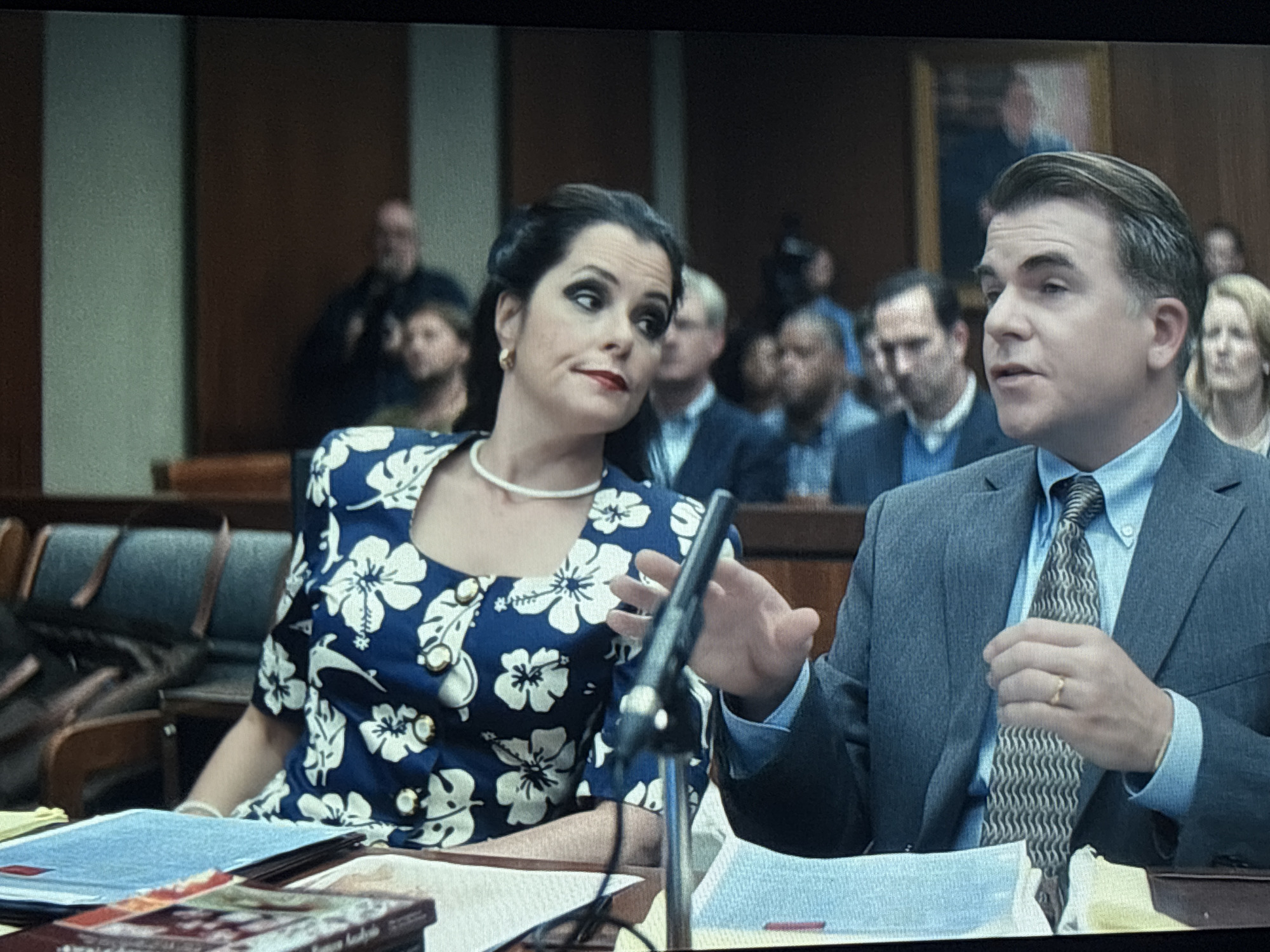 The image shows two people at a table in what appears to be a courtroom. The woman on the left is wearing a blue dress with a white floral pattern and a white pearl necklace. She has dark hair pulled back and is looking towards the right. The man on the right is wearing a gray suit, a light blue shirt, and a patterned tie. He is gesturing with his right hand as if speaking. A microphone stands between them. In the background, there are rows of people seated in the gallery. The environment is typical of a courtroom, with wood paneling and benches.
There is no information in the image that points to the city of Buenos Aires, Argentina.