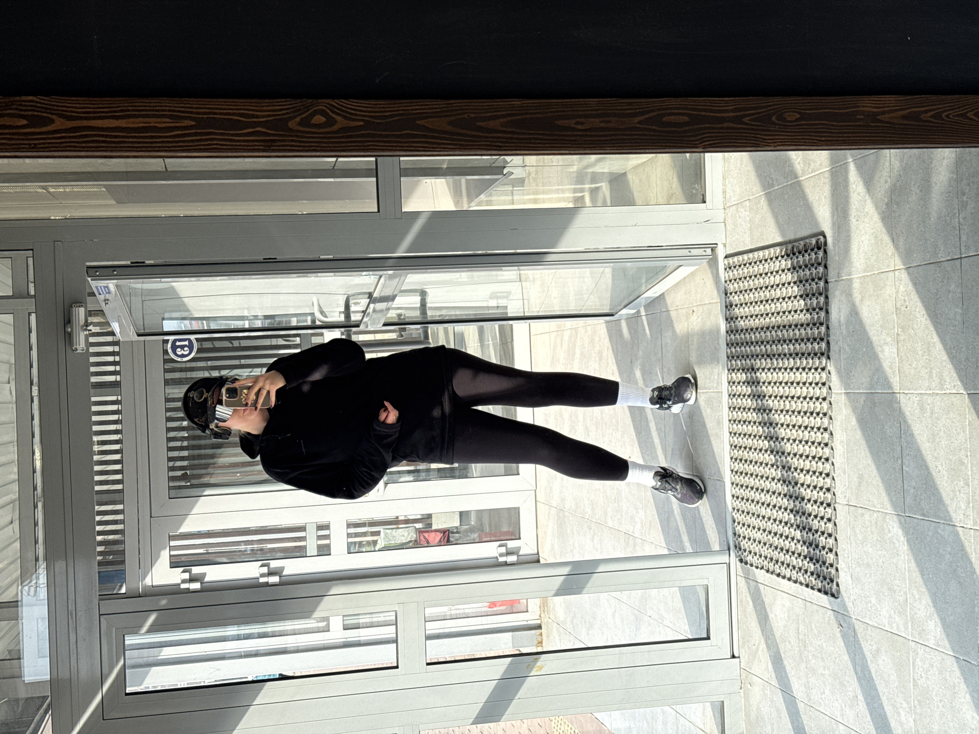 A person wearing black athletic wear, white socks, and dark sneakers stands inside a doorway in Krasnoyarsk, Russia. The doorway is made of glass and metal and includes a gray mat in front of it. The person is holding a cell phone up to take a selfie. The location appears to be indoors, with tile flooring and a wooden beam visible above the doorway. Light is streaming through the glass, creating shadows on the floor. A sign with the number 13 is visible on the glass door.