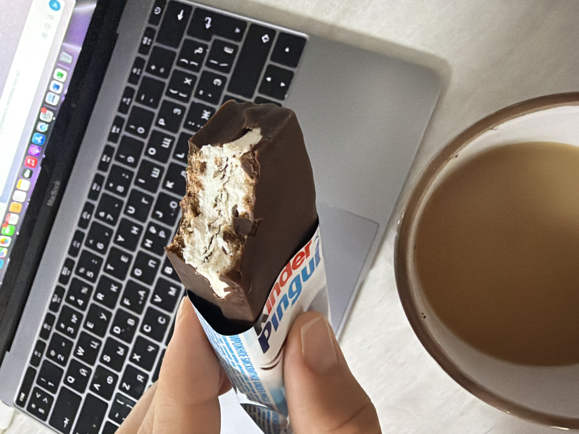 A close-up shot shows a hand holding a half-eaten Kinder Pinguí chocolate snack bar in front of an open laptop computer. The snack bar has a dark chocolate coating and a white cream filling with sponge cake. The laptop keyboard, with Cyrillic characters, is visible. In the lower right corner, a clear glass of brown liquid, likely coffee or tea, is partially visible. The background appears to be a plain white surface. There is no identifiable location in Chaikovsky, Russia.