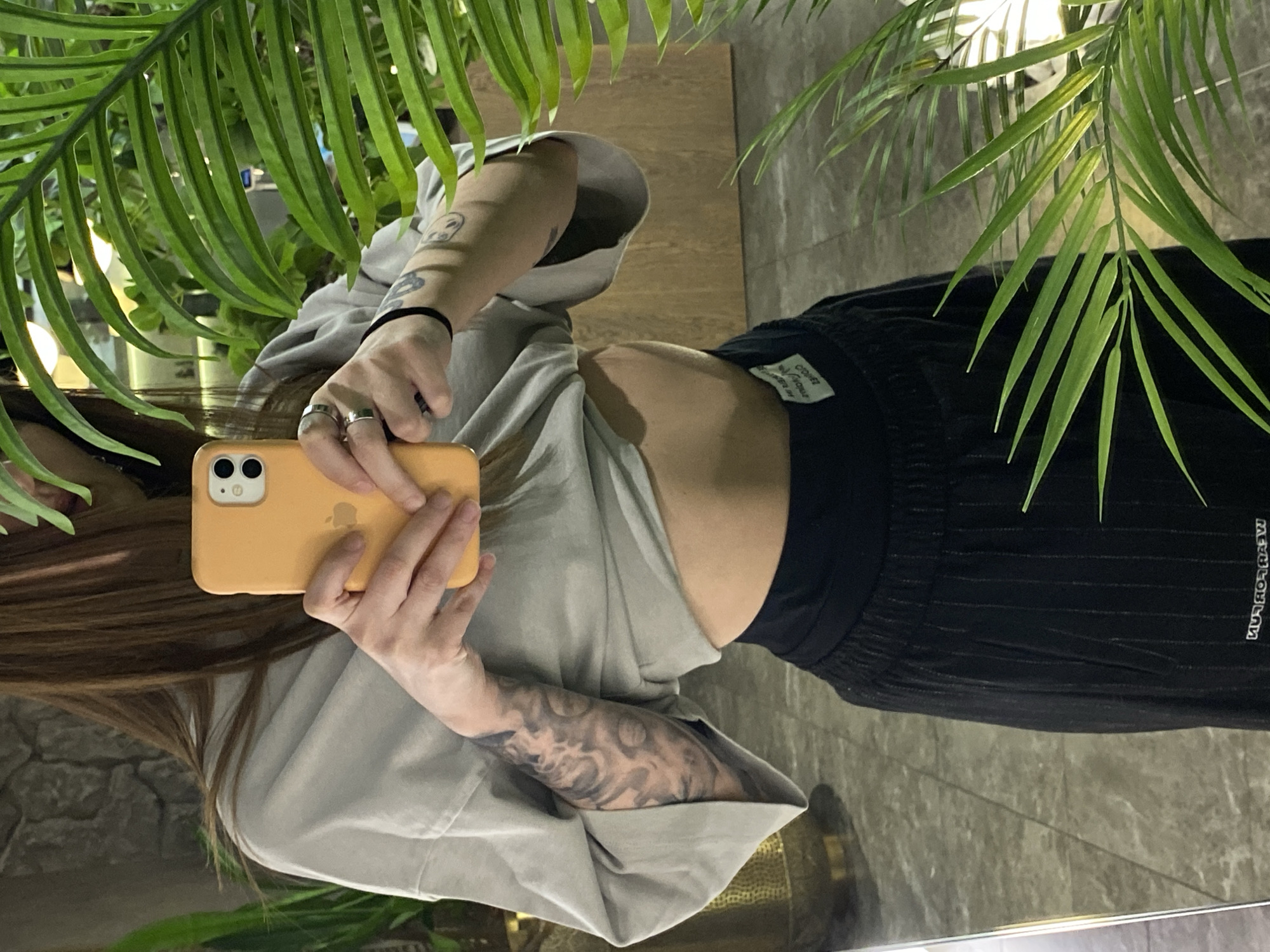 A woman is taking a selfie in a location with tropical foliage. The woman is wearing a grey t-shirt and black pants. Her left arm is visible, showing a detailed tattoo. She is holding an orange smartphone. The background includes flooring and a wall. The location is ambiguous and the relationship to Votkinsk, Russia is not evident.