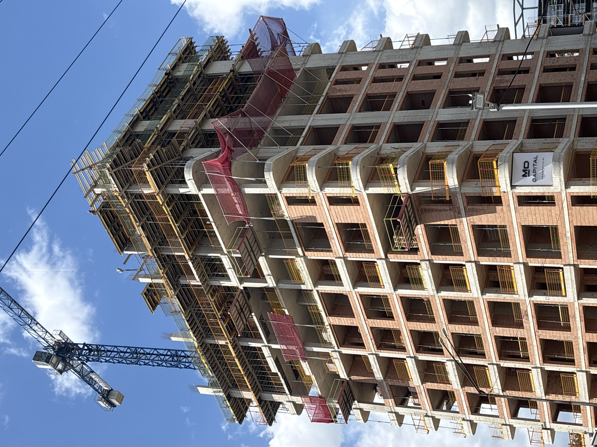 The image shows a high-rise building under construction against a bright blue sky with scattered clouds. The building's skeletal structure is visible, with exposed concrete and brickwork forming a grid of window openings. Some openings feature yellow safety cages. Construction scaffolding, covered in some areas with red safety netting, surrounds the upper floors. A construction crane is visible in the lower left portion of the frame. A sign with "MD CAPITAL" is attached to the building. The scene is in Sofia, Bulgaria.