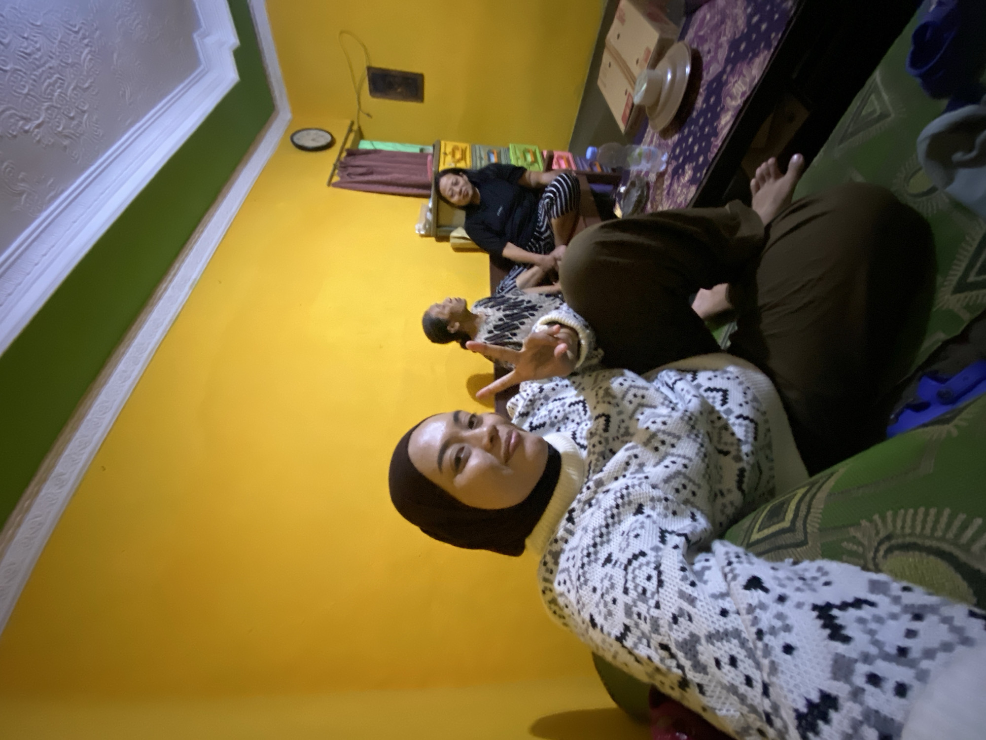 In the city of Kecamatan Pati, Indonesia, a group of individuals is gathered in a room. The dominant color of the walls is a vibrant yellow, with a green band across the ceiling, which is ornately decorated. In the foreground, a person wearing a dark hijab and a white and grey patterned sweater is seated on a sofa, looking towards the camera and holding up two fingers in a peace sign. To their right, two other individuals are seated. One, wearing a black and white patterned top, is looking to their left, while the other, in a black t-shirt and striped skirt, is looking forward. A small table with a purple cloth and various objects, including a bottle of water, a box, and a hat-like object, is positioned in front of them. A clock is visible on the wall in the background, near a window with dark curtains. A stack of colorful drawers also appears to be part of the room's furnishings. The overall impression is an indoor social gathering.