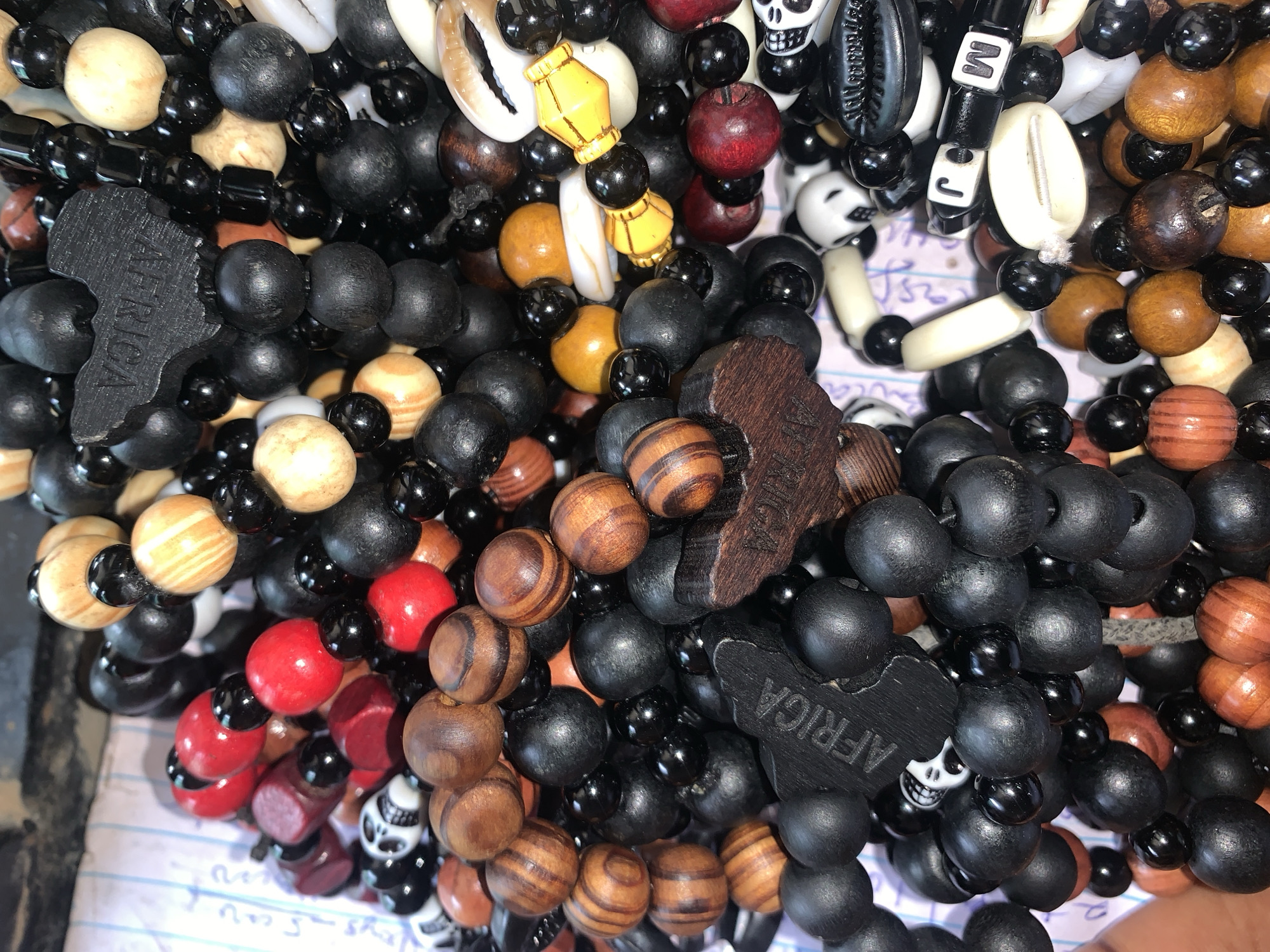 A close-up shot shows a collection of beaded bracelets. The bracelets feature a variety of bead types, including wooden beads in various shades of brown, black beads, red beads, and light yellow beads. Some beads are shaped like skulls. Several bracelets have a charm in the shape of the continent of Africa. Other bracelets include cowrie shells. The bracelets are piled together. The setting appears to be indoors, possibly in Kabong, Nigeria. The focus is on the bracelets themselves, highlighting their diverse materials and design.