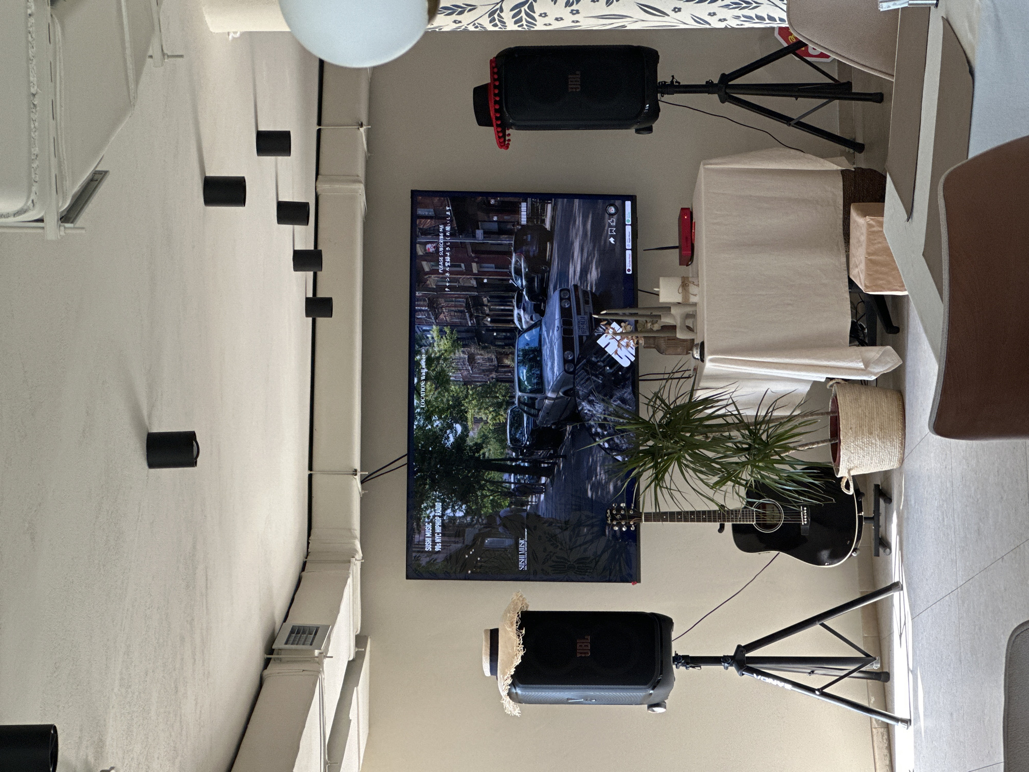 The scene shows the interior of a room with a television mounted on a wall, displaying a video about the city of Boreti, Montenegro. The video shows city streets with a car with a license plate labeled "XFP-9434". Beneath the video text reads "PLEASE SUBSCRIBE ME ・ネル登録よろしくお願い" on the left side of the screen and some social media icons in the upper right corner. Below the television, to the left and right are JBL speakers mounted on stands, with a guitar and potted plant placed in front of the speaker to the right. The room is decorated with multiple cylindrical light fixtures along the ceiling's edge. A table and chair are on the right side of the scene.