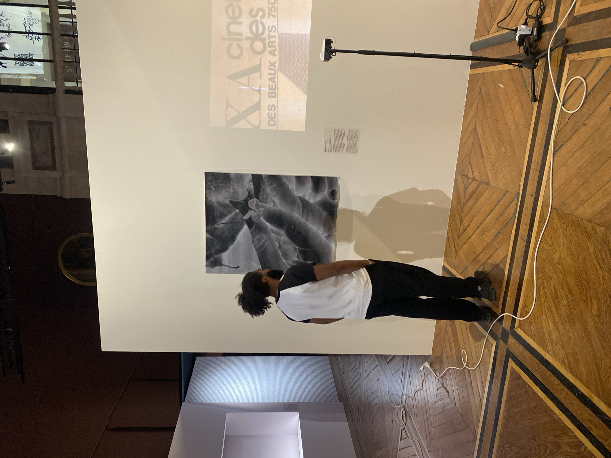 The image depicts a person looking at an art installation on a wall. The individual is wearing a white and black shirt with dark pants and shoes. The wall displays a black and white photograph, a text panel, and a projected graphic that reads "XA cinema des BEAUX ARTS 750". A black camera stand is positioned to the right of the installation. The flooring consists of a wooden parquet pattern with dark inlays. Additional art exhibition structures are positioned at the bottom of the image. The location is plausibly in Paris, France, based on the text in the image.