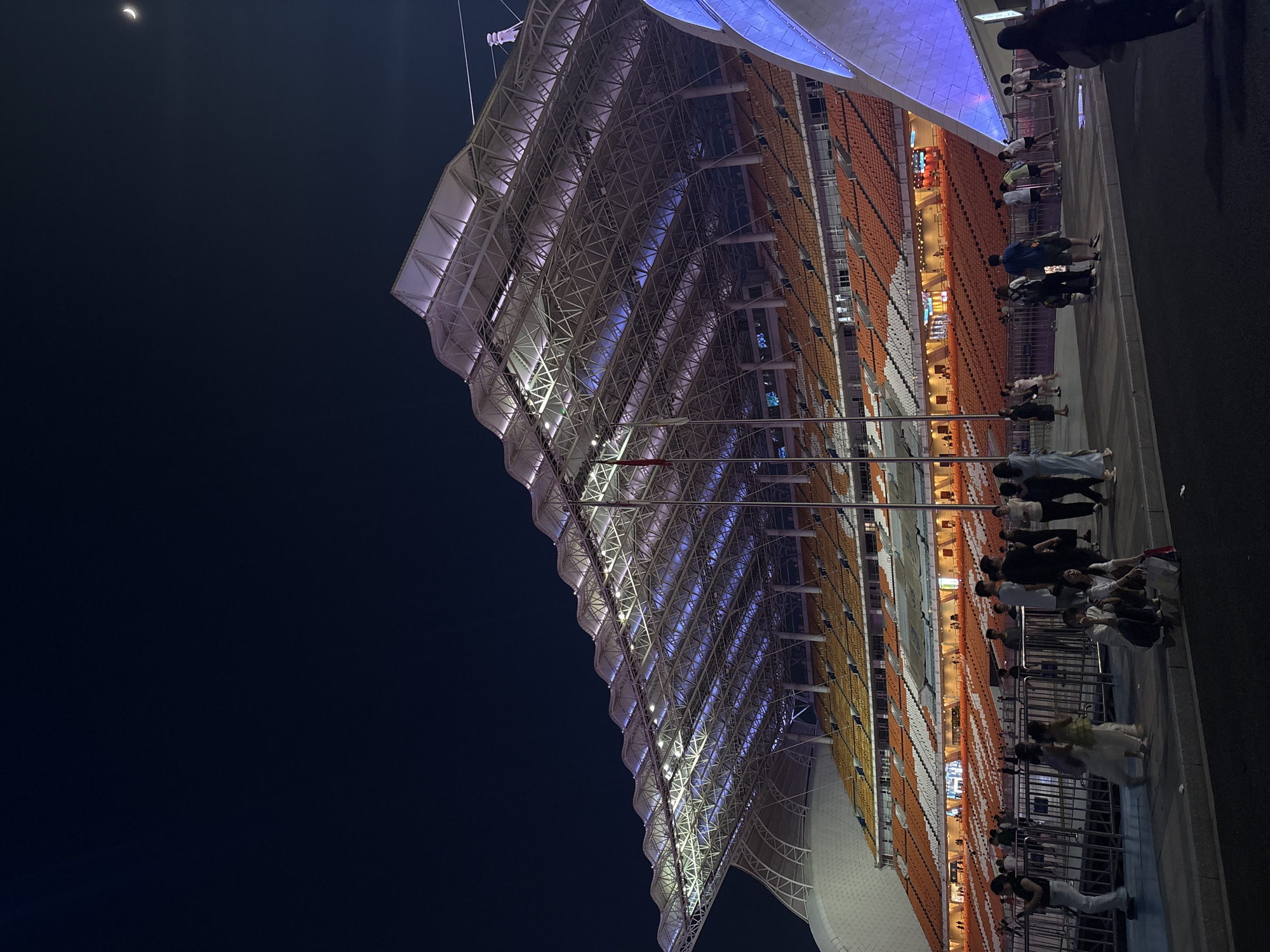 The image shows a large stadium at night. The stadium has a distinctive roof structure with a metallic framework illuminated by white and blue lights. The seating areas are visible, with rows of orange and white seats. People are gathered outside the stadium, walking along a pavement next to a metal barrier. Some individuals are standing in small groups, while others are walking individually. The sky is dark, indicating it is nighttime. A sliver of the moon is visible in the upper left corner. The lighting from the stadium creates a vibrant atmosphere. The scene appears to be a public gathering, possibly before or after an event at the stadium. The location cues suggest it is in Tianhe District, China.