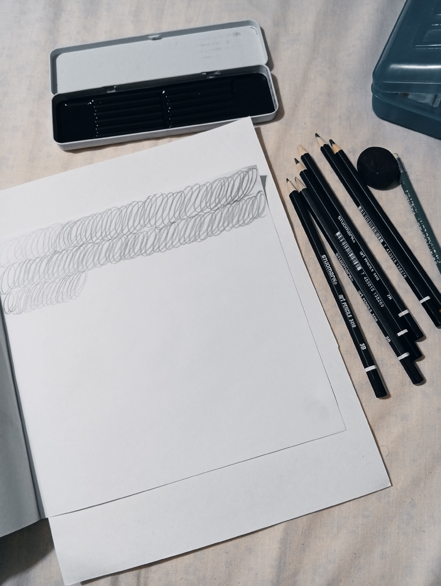 A high-angle medium shot captures a flat lay of art supplies on a light-colored textured surface, likely a tablecloth or fabric. The scene is dimly lit, with a cool, muted color palette. In the upper left corner, a rectangular silver tin box is open, revealing a collection of black graphite drawing pencils. To the right of the tin, several black graphite pencils are scattered on the surface, some with visible "STUDIOMARK Art PENCILS 3410" and hardness indicators like "7R" and "3B" printed on them. An eraser is also visible near the pencils.
In the foreground, a white sheet of paper lies open, with a sketch of two rows of overlapping, loosely drawn ovals or loops in graphite. Below this paper is another white sheet of paper. The background is a textured fabric surface. The image does not contain any elements that suggest a specific location in San Antonio de Los Altos, Venezuela.