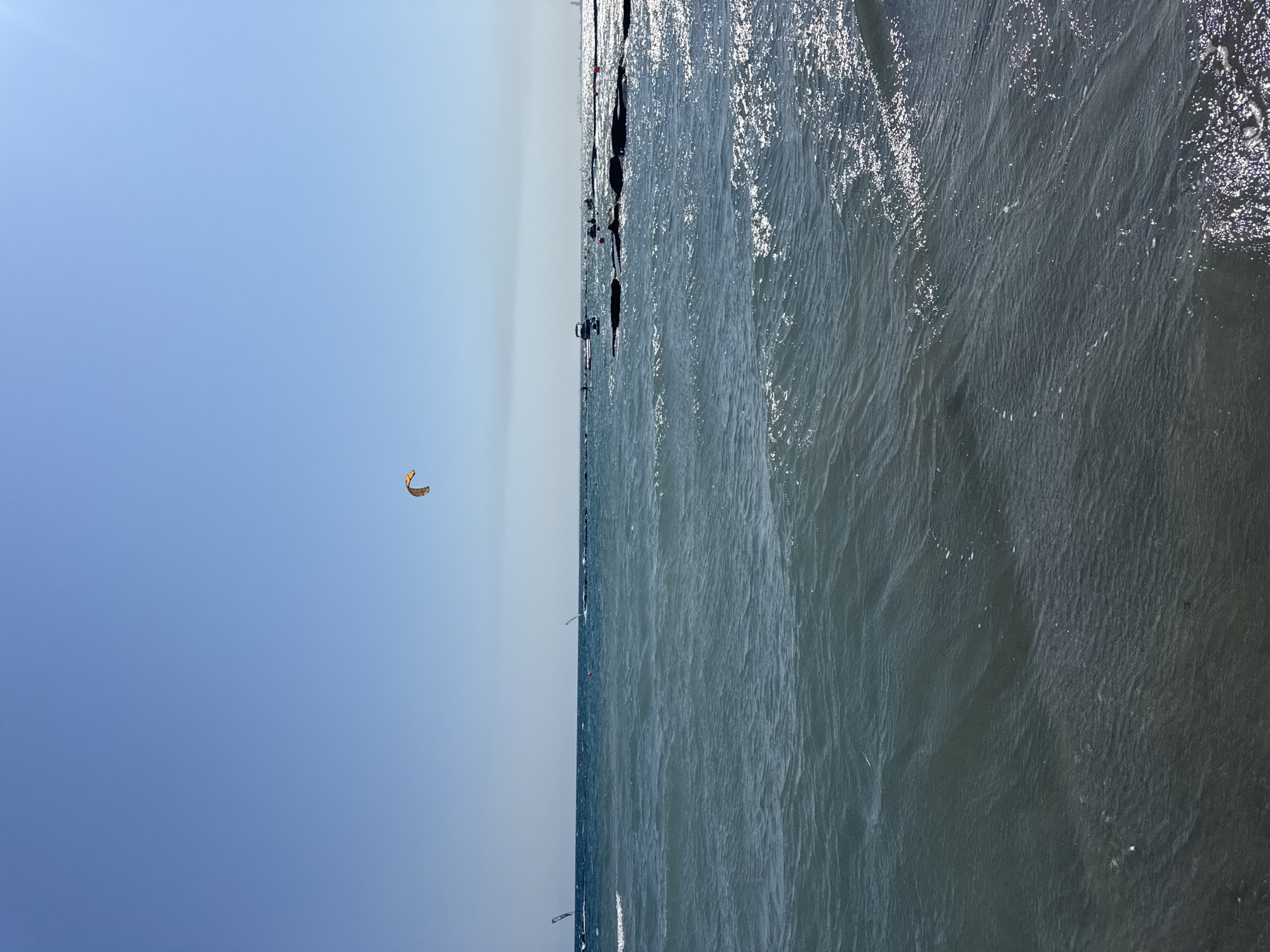 The image depicts a sunny day at the beach in Agios Tychona Community, Cyprus. The clear blue sky is visible above the calm, blue sea. The water has gentle waves, and the sunlight creates a shimmering effect on the surface. In the distance, several kite surfers are enjoying the activity, with their colorful kites visible against the sky. There are also some boats and buoys in the water, indicating a recreational area. The overall scene is one of a peaceful and active beach day in a Mediterranean setting.