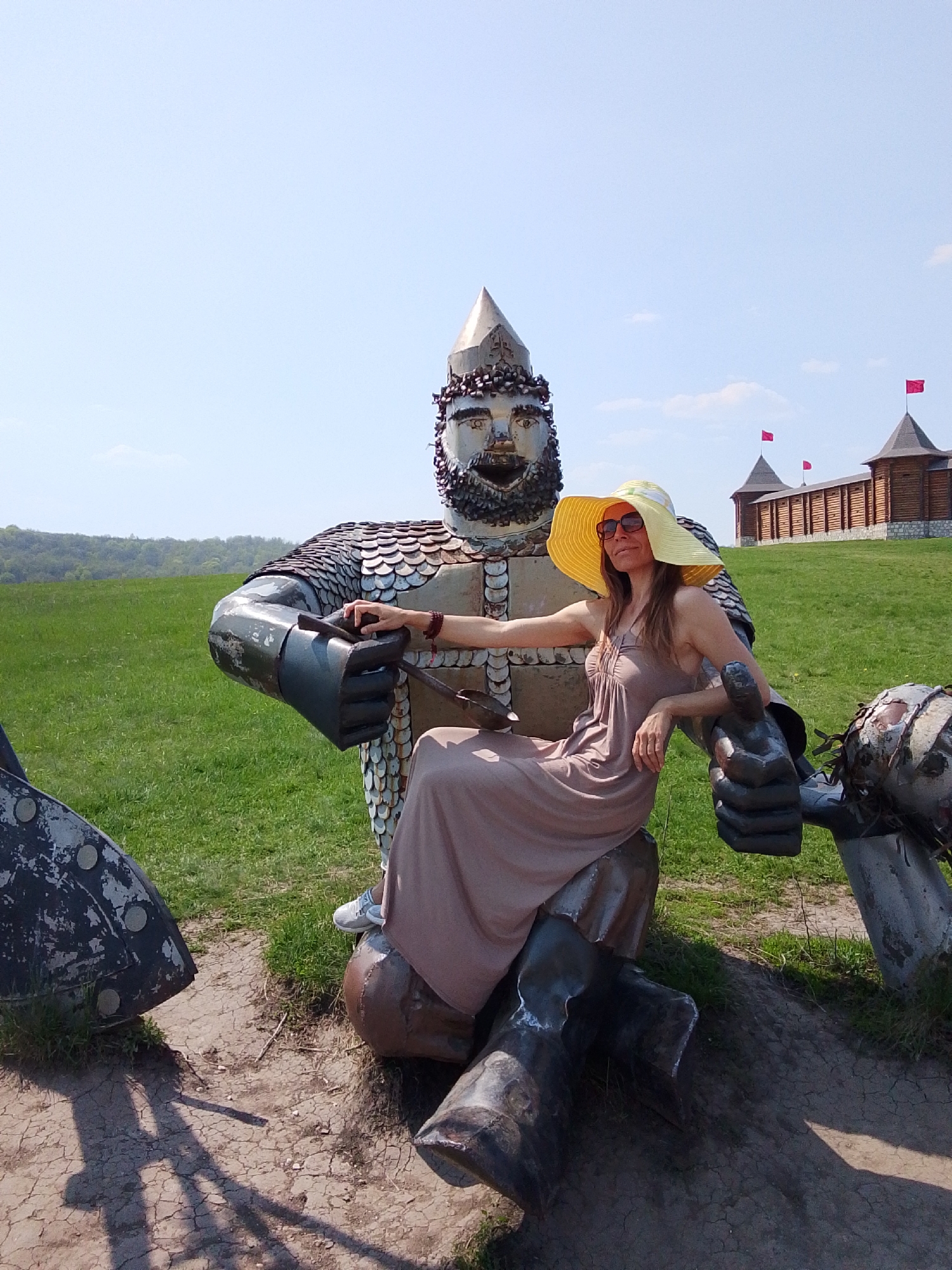 A woman wearing a yellow sun hat and a beige dress is sitting on a metal sculpture of a warrior in Каменка, Russia. The warrior sculpture has a helmet and is covered in metal scales. In the background is a wooden structure with red flags on top. The ground is covered in grass and dirt.