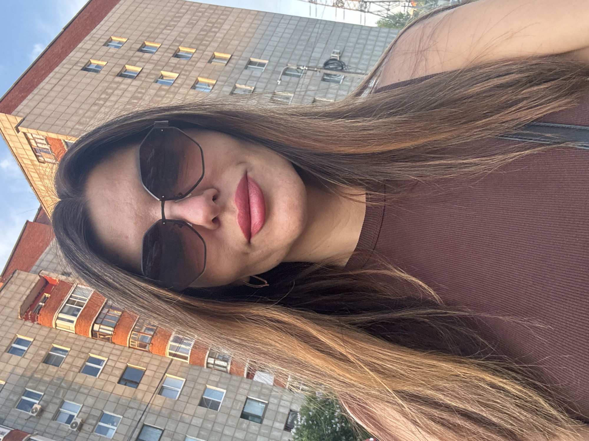 The image depicts a woman wearing sunglasses and a brown top, posing for a selfie. Her hair is long and brown, with some lighter highlights. The background features two buildings, one appearing to be gray with red brick accents and many windows, and the other a taller, more modern gray building. A construction crane can be seen in the distance. Based on the architectural style of the buildings, the picture was taken in the city of Chaikovsky, Russia.