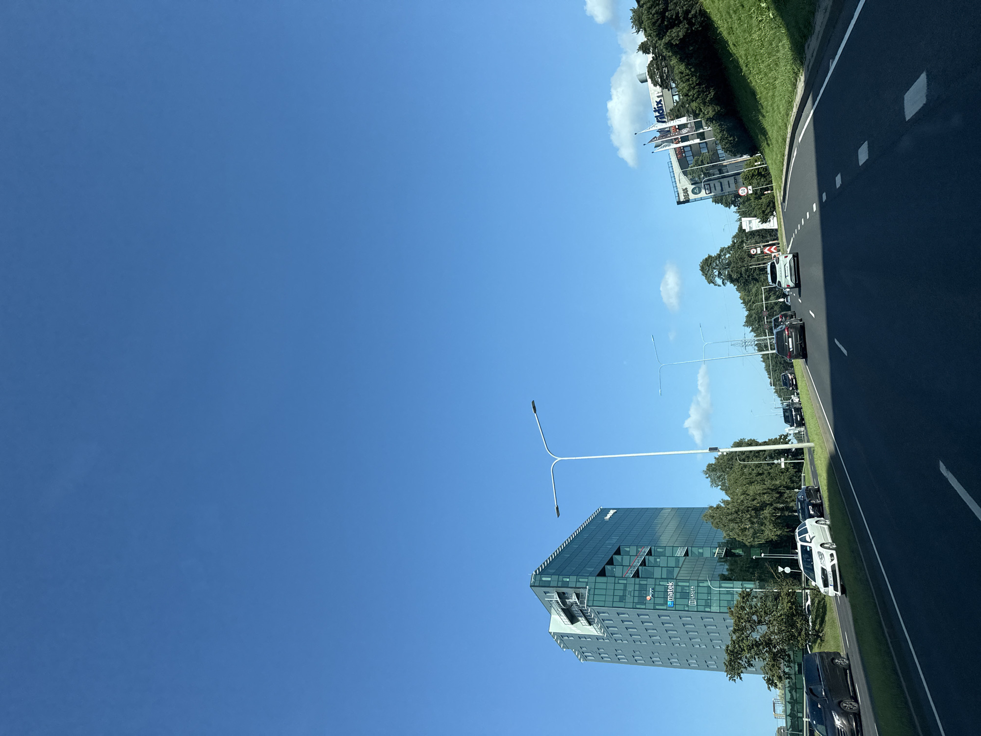 The image depicts a clear, sunny day in Tallinn, Estonia, with a bright blue sky. The scene is a multi-lane road, likely an urban arterial, with cars in motion. On the left side of the road stands a modern, multi-story office building with a glass facade, featuring green tinted windows and visible architectural details like external staircases. The building has signage, including "matek" and "LASITA". Opposite this building, on the right side of the road, is a grassy embankment leading up to a lower structure with several flags. This building also has signage, including "vidix" and "IDEAS". Streetlights and traffic signals are visible along the roadside. The road itself is asphalt with clear lane markings. The overall impression is of a busy, modern commercial or business district.