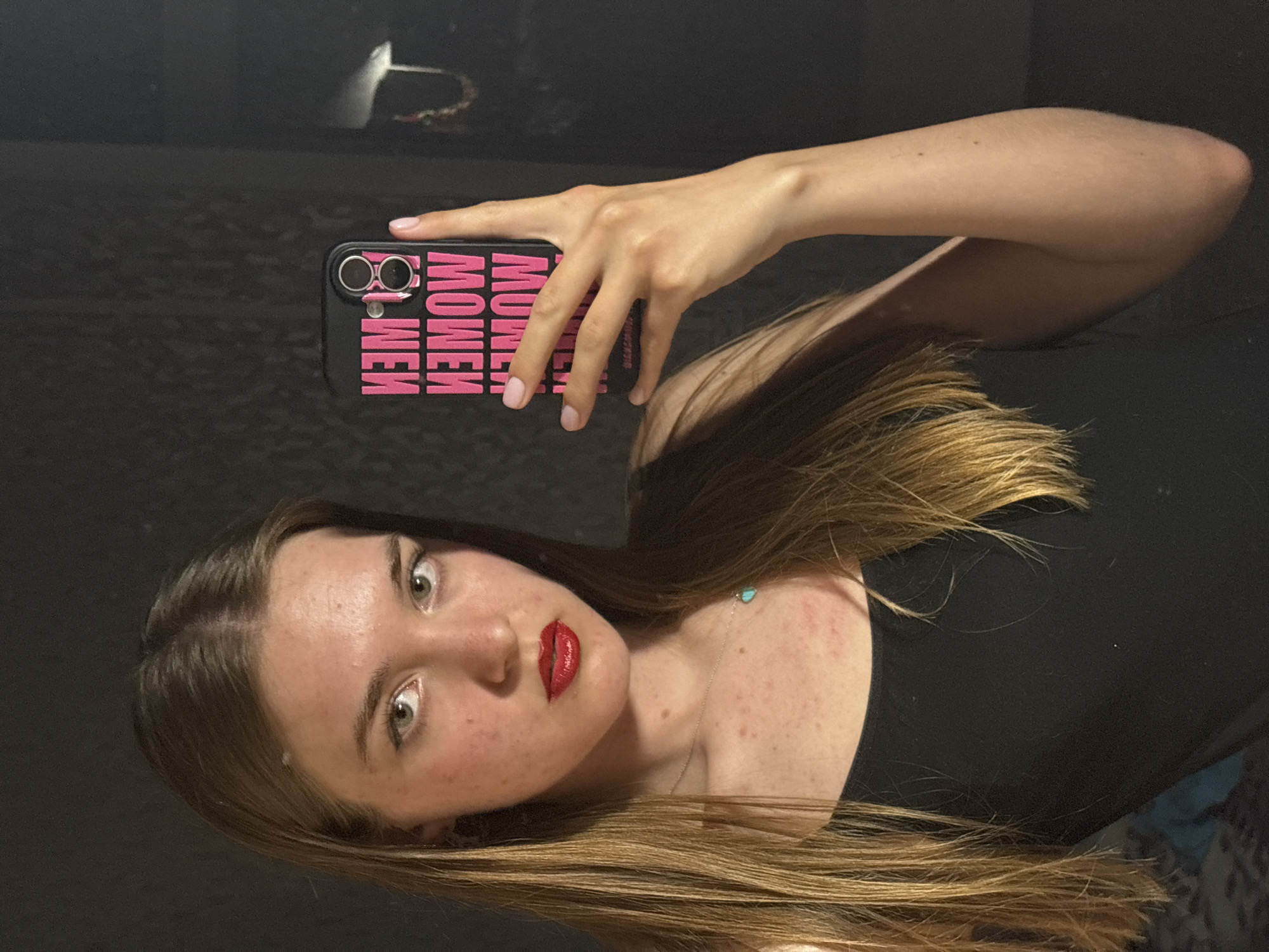 A fair-skinned young woman with light brown hair and red lipstick is taking a selfie using a smartphone in a black phone case with the word "WOMEN" printed repeatedly in pink. She is wearing a black top and a silver necklace with a small turquoise pendant. The background is dark and indistinct, suggesting an indoor setting. The location is not discernible from the image.